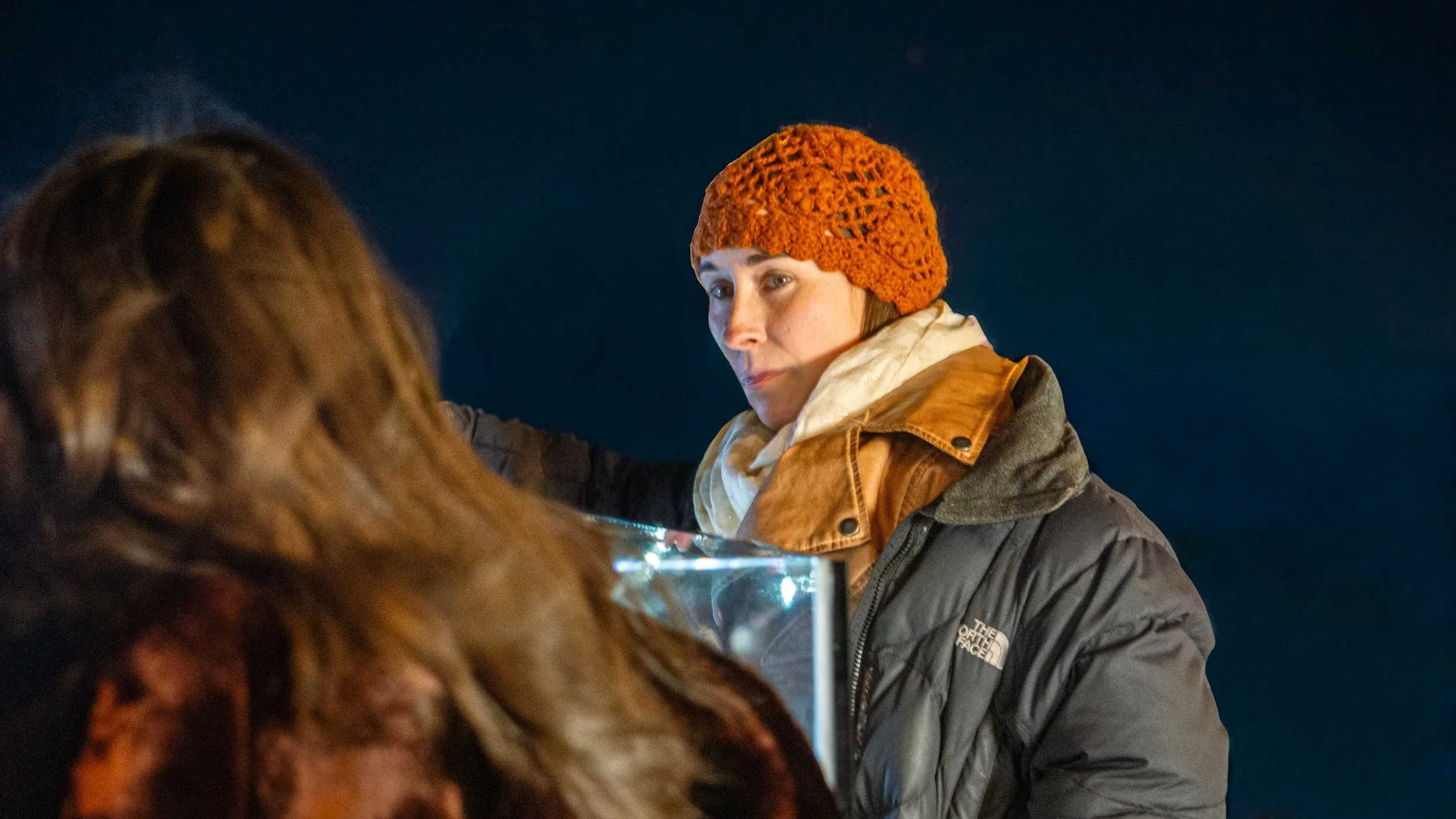 Person with short dark hair wearing an orange knit hat and a brown North Face jacket at night, looking at a display.