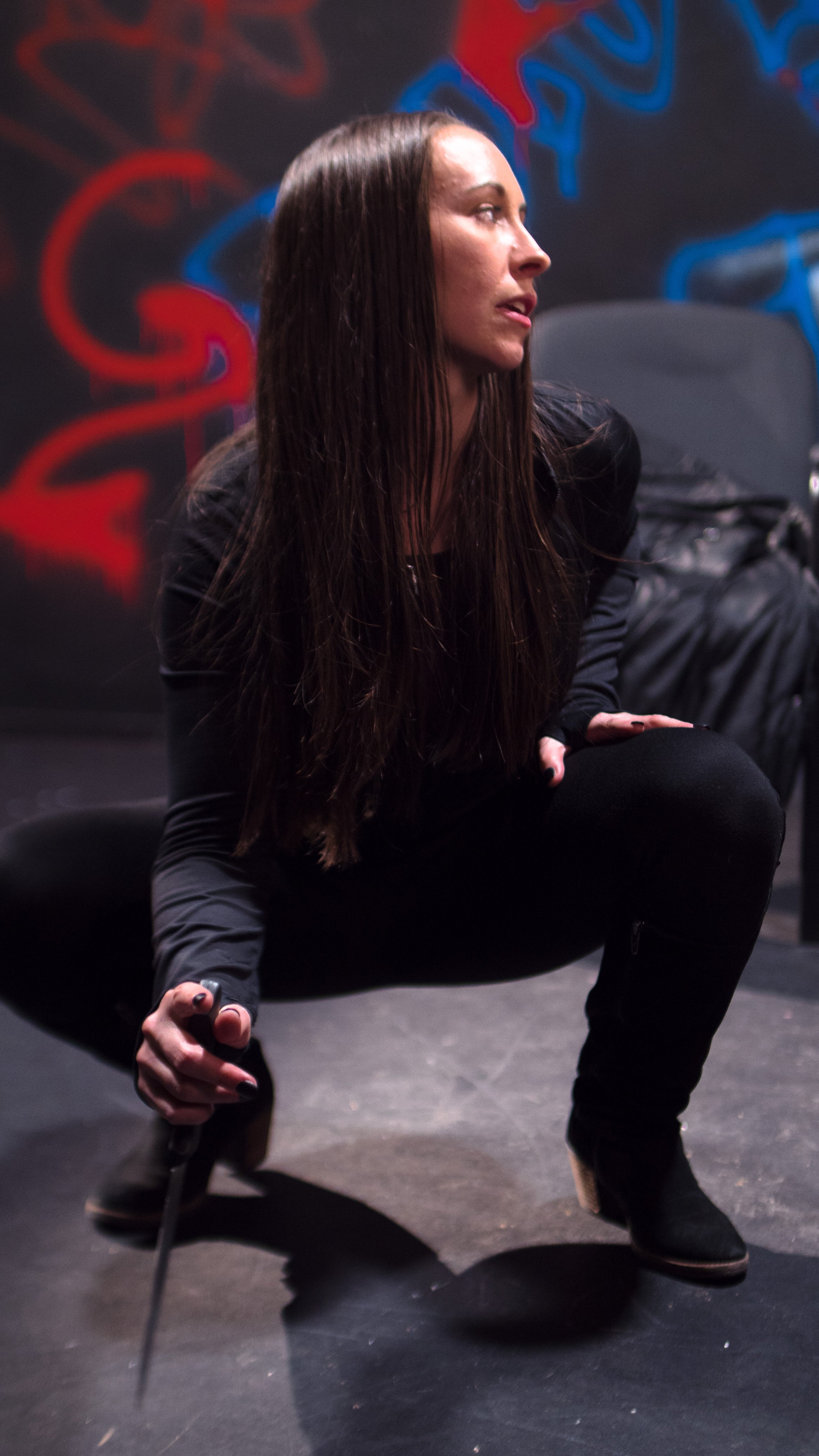 Woman with long dark hair in a crouching pose, wearing black clothing and boots, on a stage or indoor setting with graffiti-style wall art in red and blue.