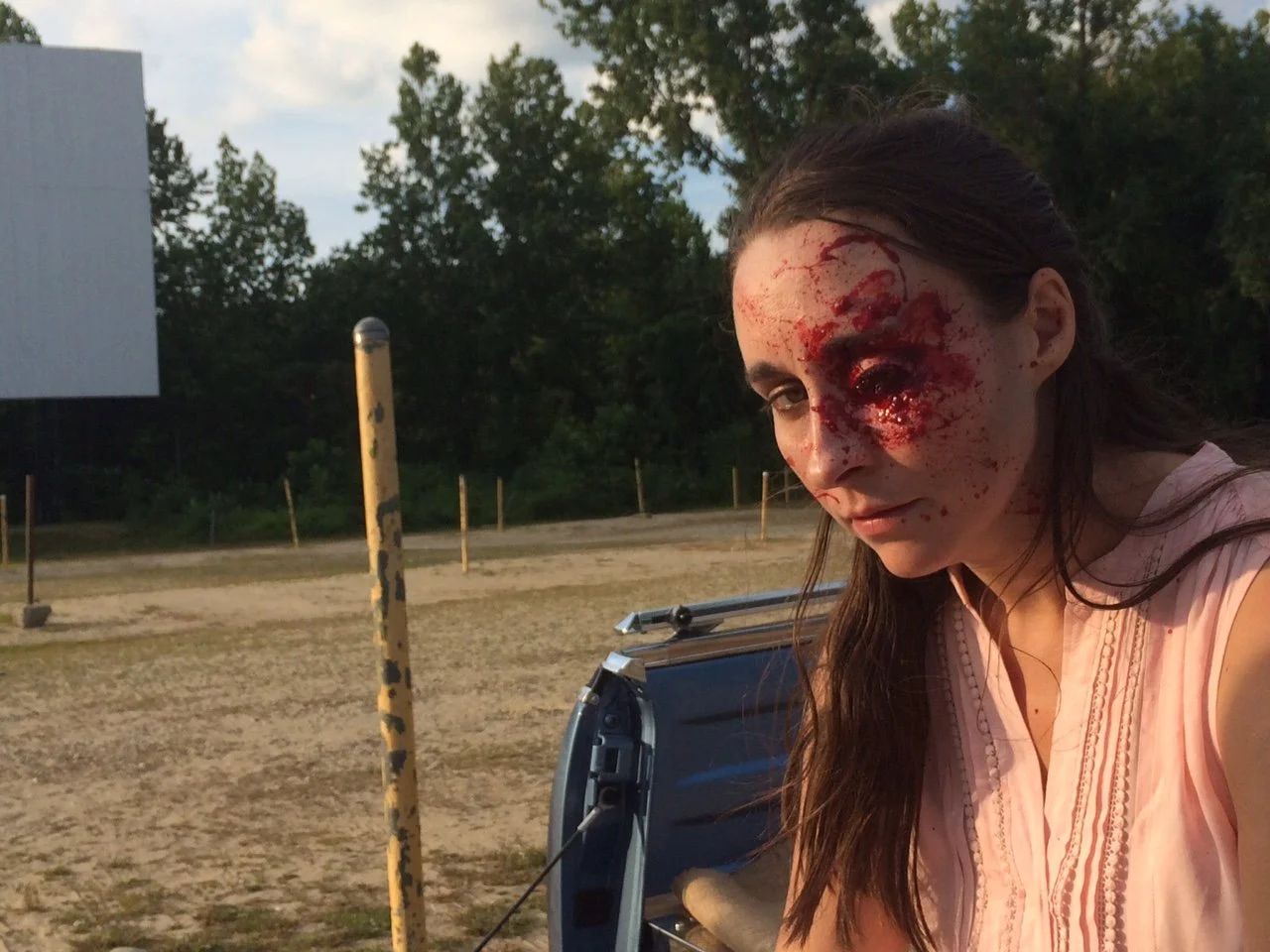 Woman with makeup or special effects makeup resembling a severe facial injury or zombie wound, outdoors in a dirt area with green trees and a cloudy sky in the background.