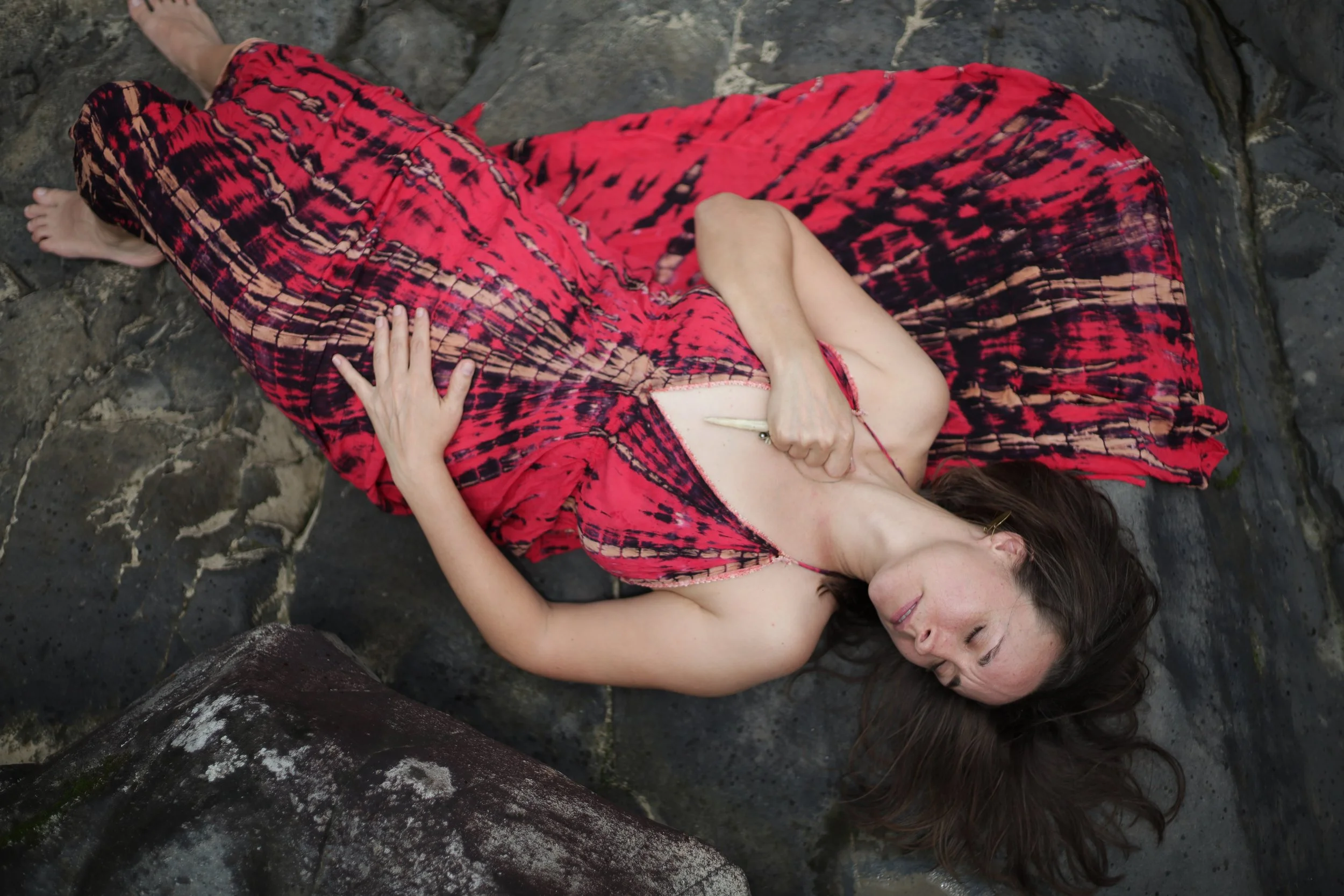 A woman in a red and black tie-dye dress lying on a rocky surface with her eyes closed and a serene expression.