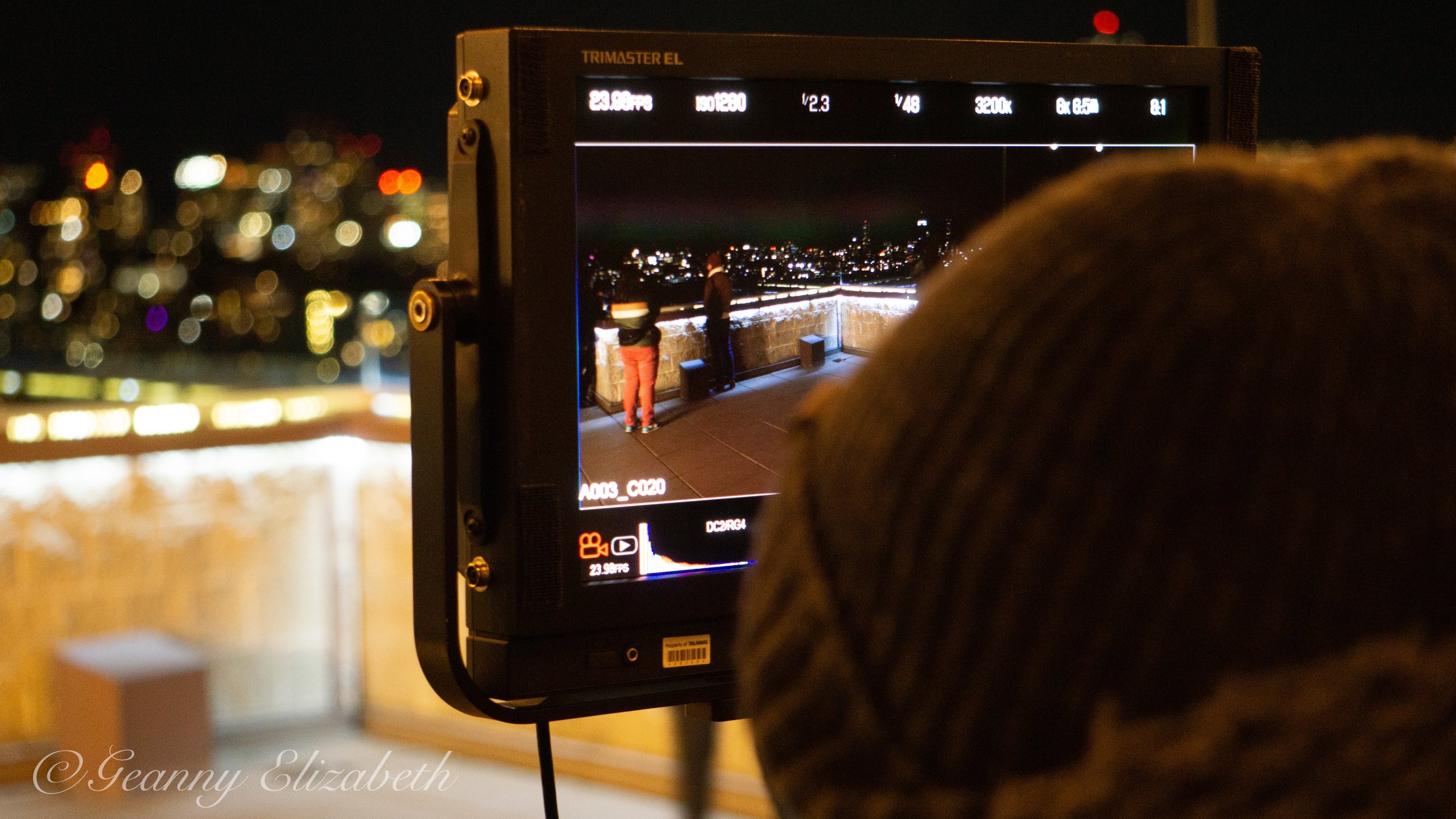 A camera monitor displaying a night scene of two people standing on a rooftop overlooking a city skyline with many blurred lights in the background.