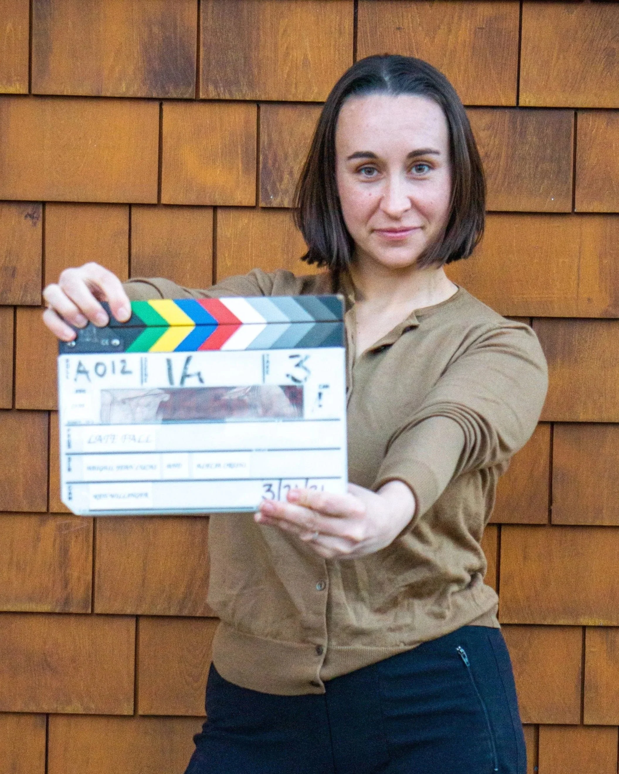A woman with dark hair and blue eyes holding a movie clapper board in front of her. She is standing in front of a wooden wall and wearing a brown long-sleeved shirt and dark pants.