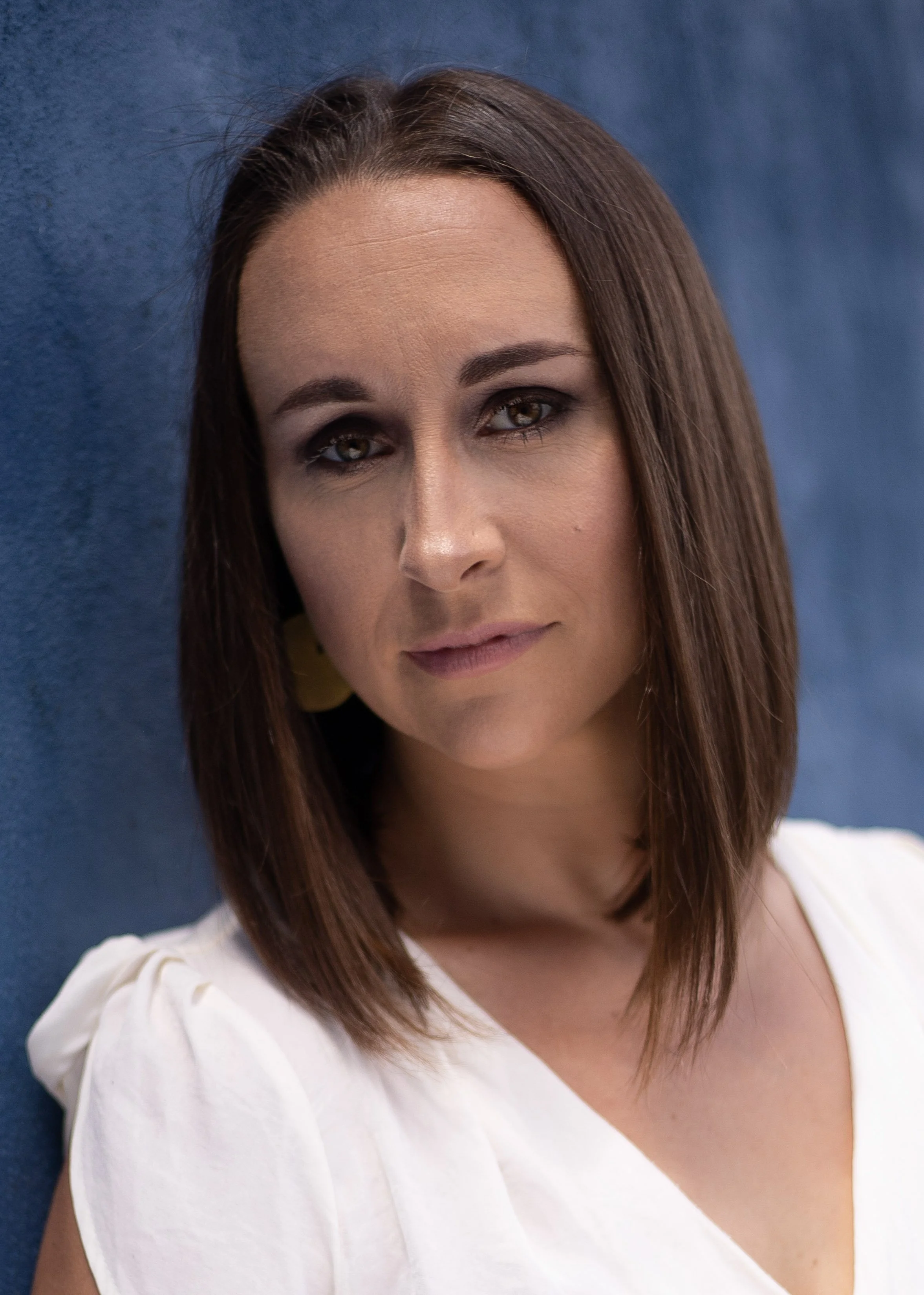 A woman with brown hair and hazel eyes wearing a white top, standing in front of a blue background.