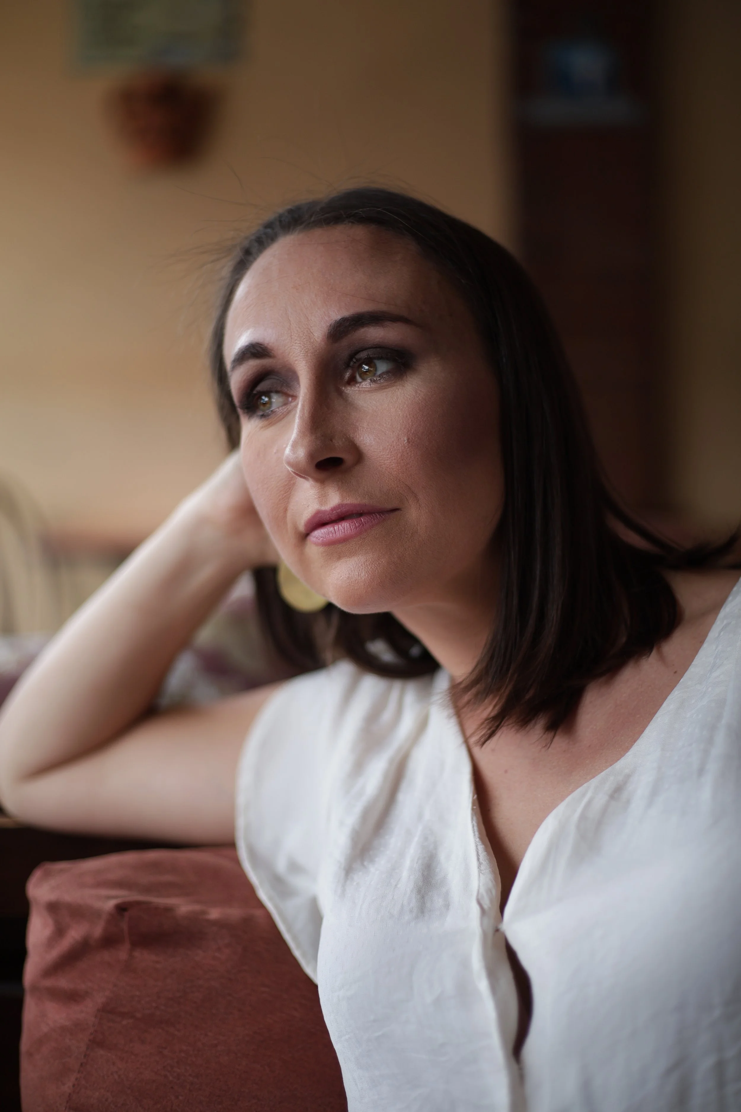 Close-up of a woman with dark hair, wearing makeup, resting her head on her hand with a relaxed expression. She is wearing a white top and is sitting on a reddish-brown couch. The background features an indoor setting with a blurred wall and some objects.