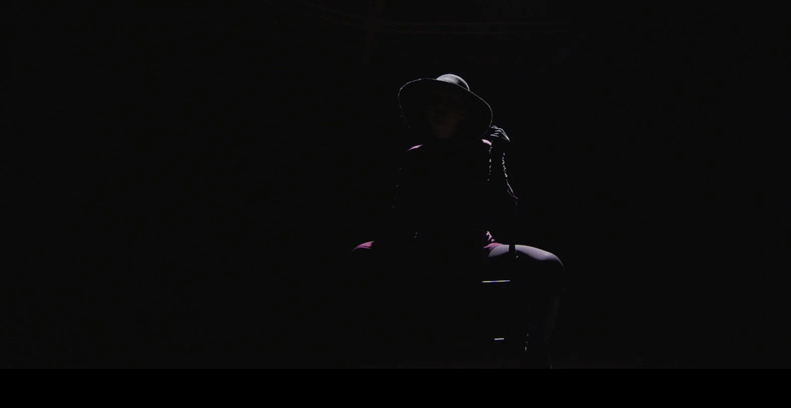 A woman wearing a large hat sitting in a dark, shadowy environment with minimal lighting highlighting her silhouette.