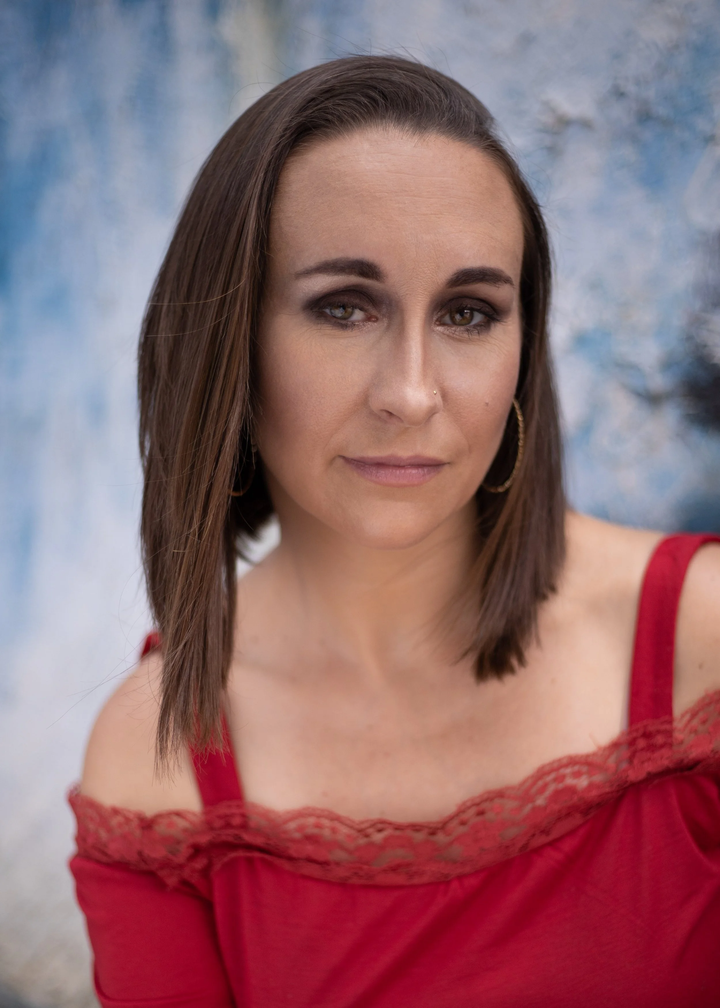 A young woman with brown hair, wearing a red dress with lace details, is looking slightly to the side with a subtle smile. The background is a blurred, abstract pattern of blue and white.
