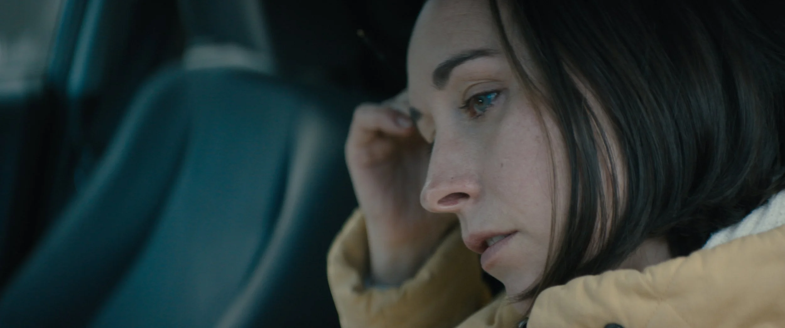 A woman with dark hair and blue eyes resting her head on her hand and looking at a device inside a vehicle.