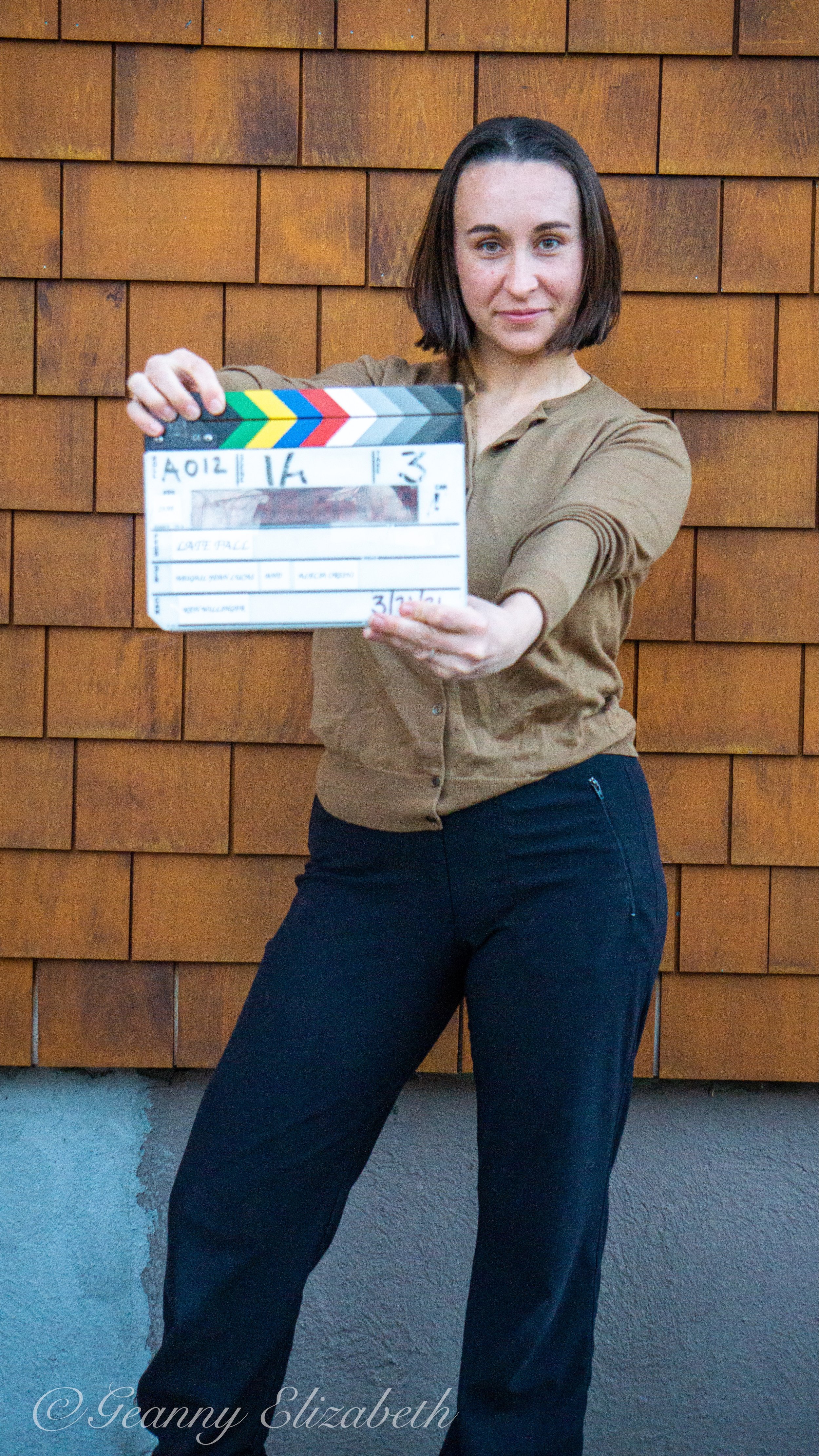 A woman holding a film clapperboard in front of a wooden wall, posing for a photo.