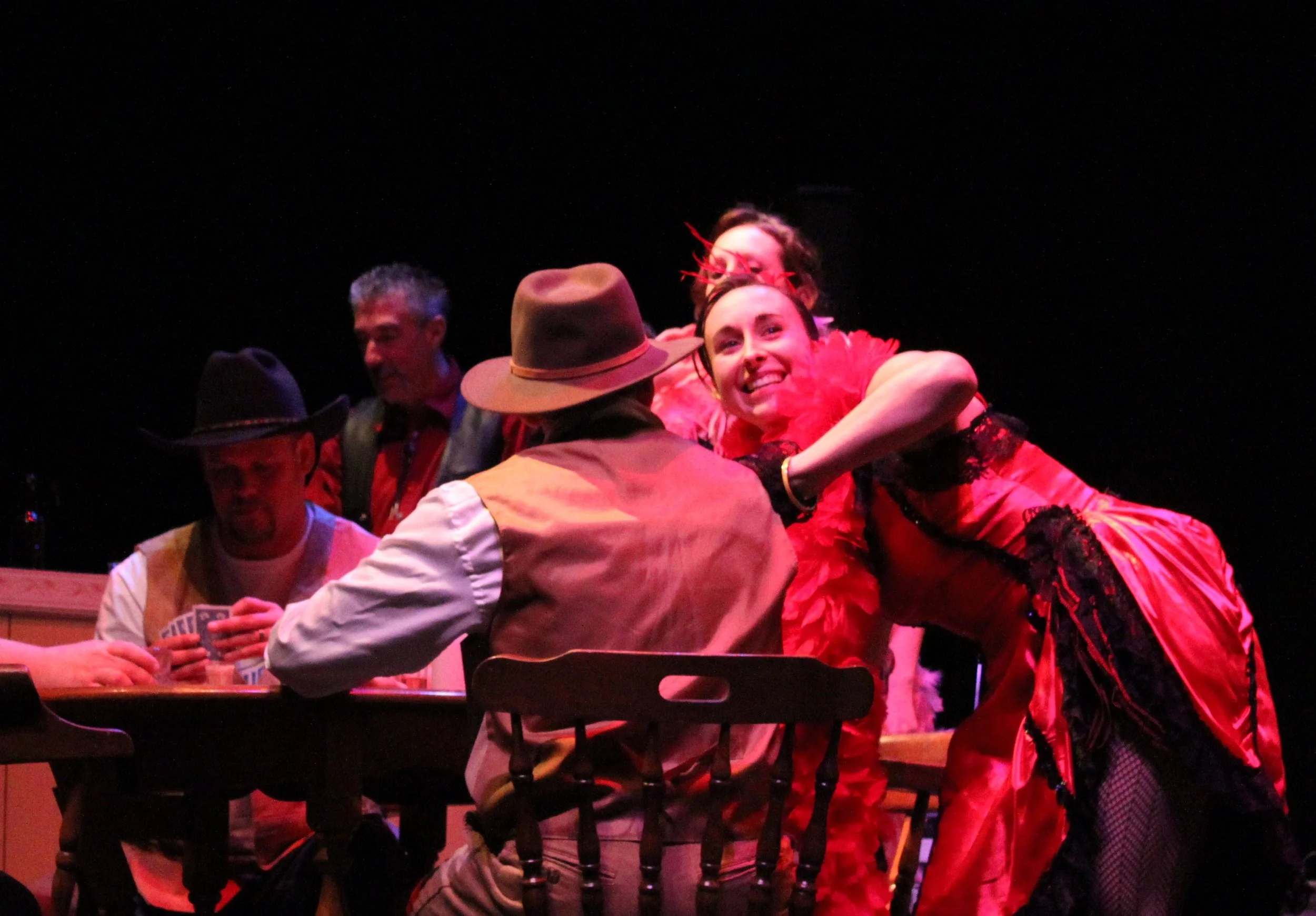 Group of people at a table, with one woman in a vibrant red dress smiling and hugging someone, others are chatting and holding drinks in a dimly lit setting.