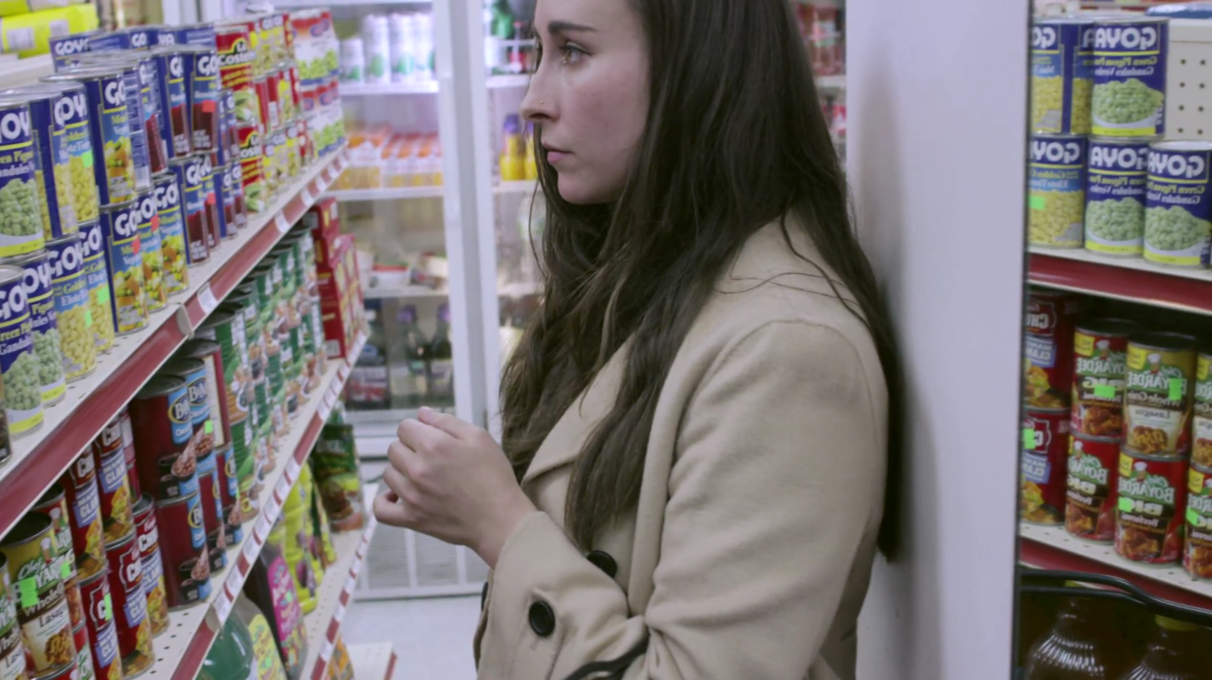 A young woman with long dark hair and a beige coat shopping in a grocery store, looking at canned goods on a shelf.
