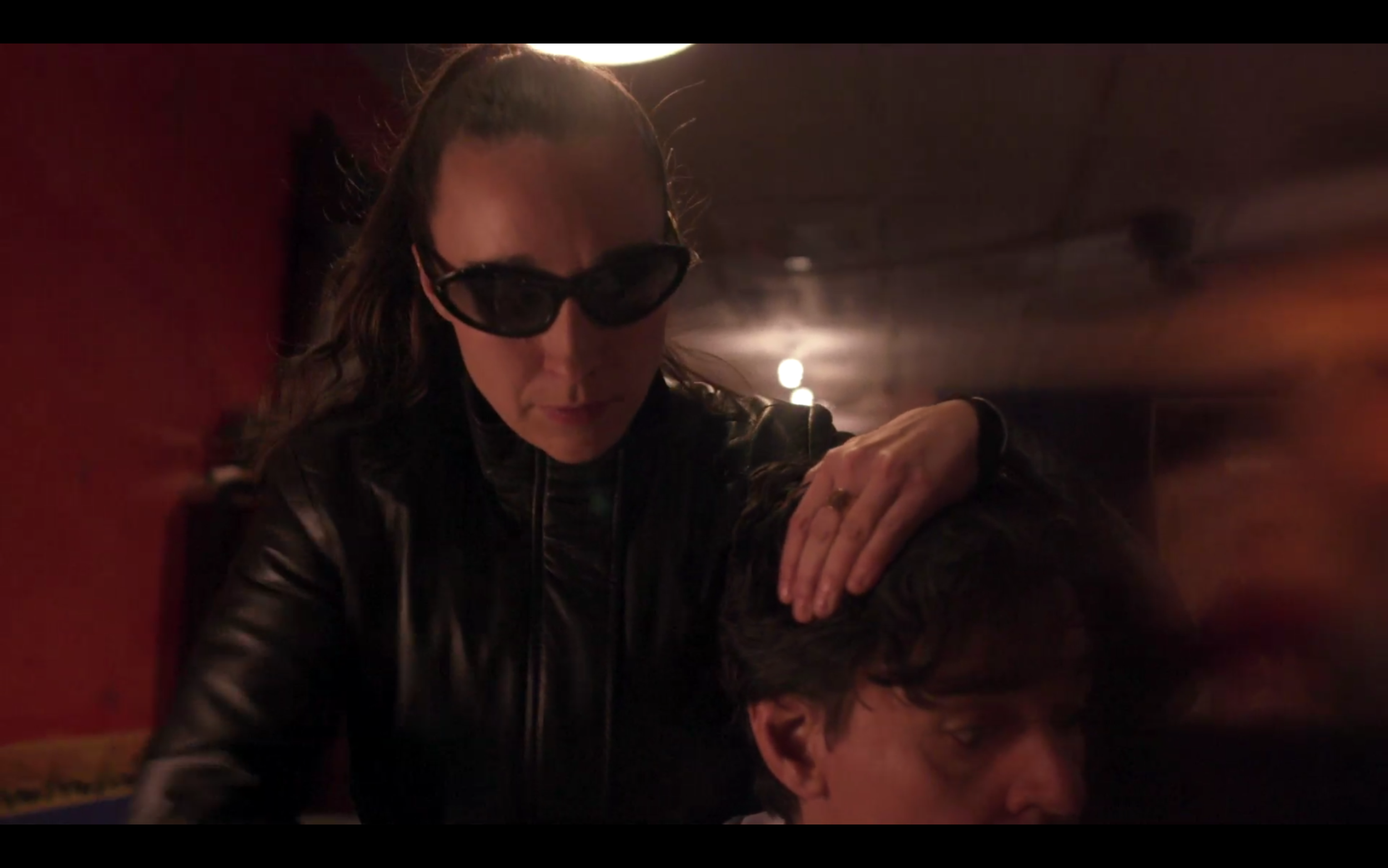 A woman with long hair in a ponytail wearing large black sunglasses and a black leather jacket, leaning over and holding the head of a man with short dark hair in front of her, inside a dimly lit room.