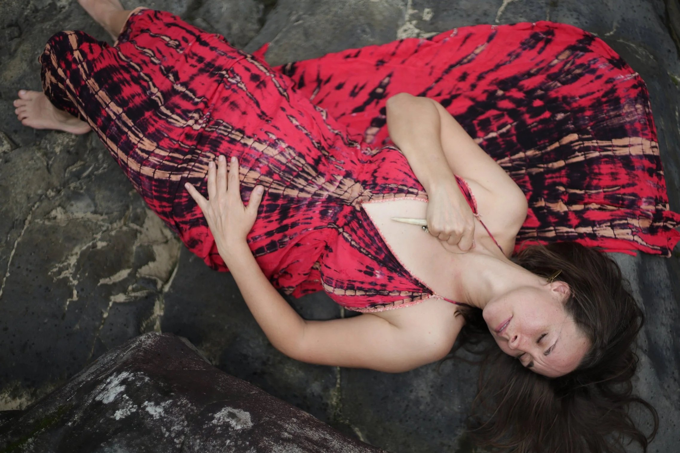 A woman with long brown hair lying on rocky ground, wearing a red and black patterned dress, with her eyes closed and arms resting on her stomach.