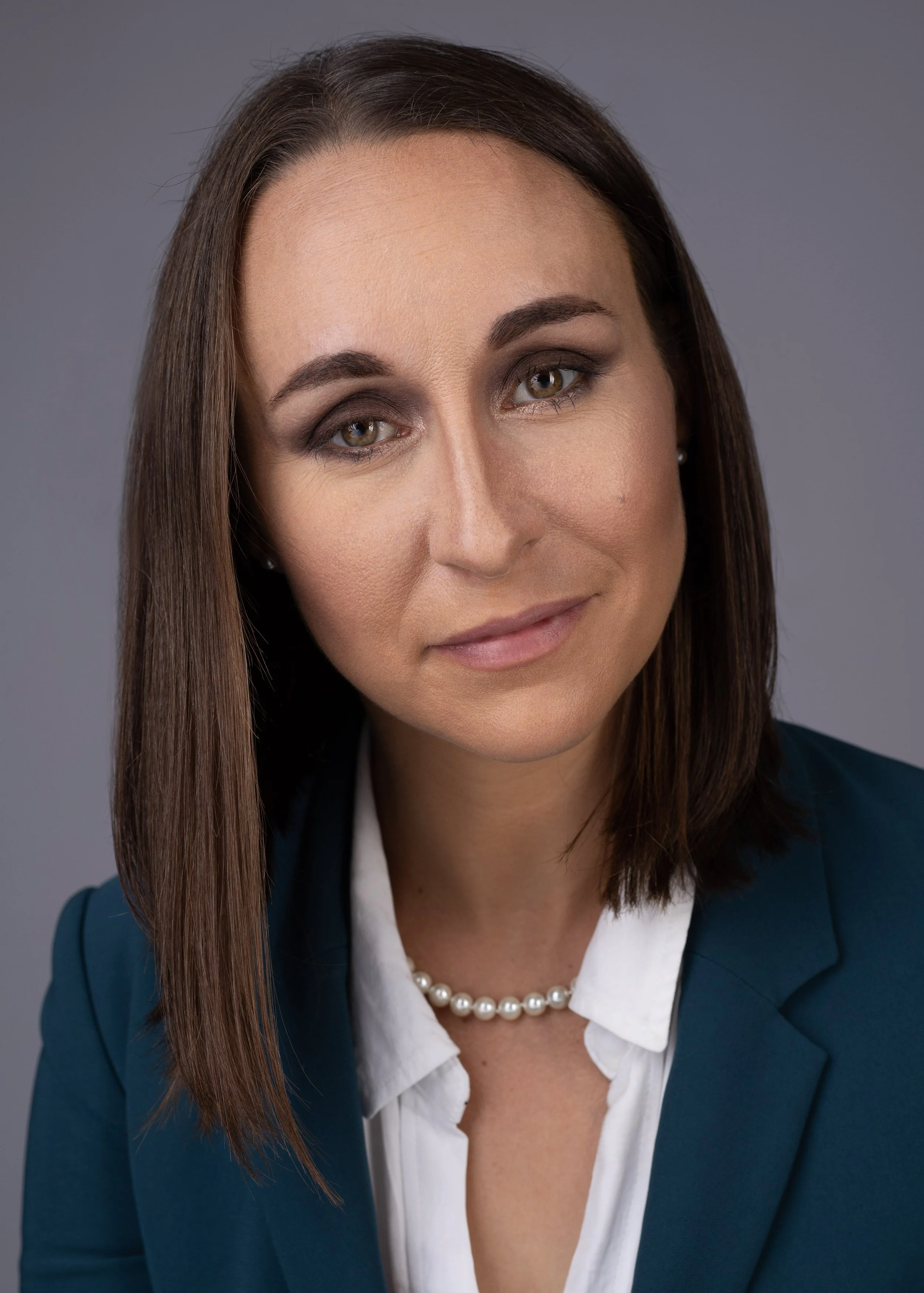 Close-up portrait of a woman with straight brown hair and hazel eyes, wearing a dark blue blazer, white shirt, a pearl necklace, and stud earrings, against a plain gray background.