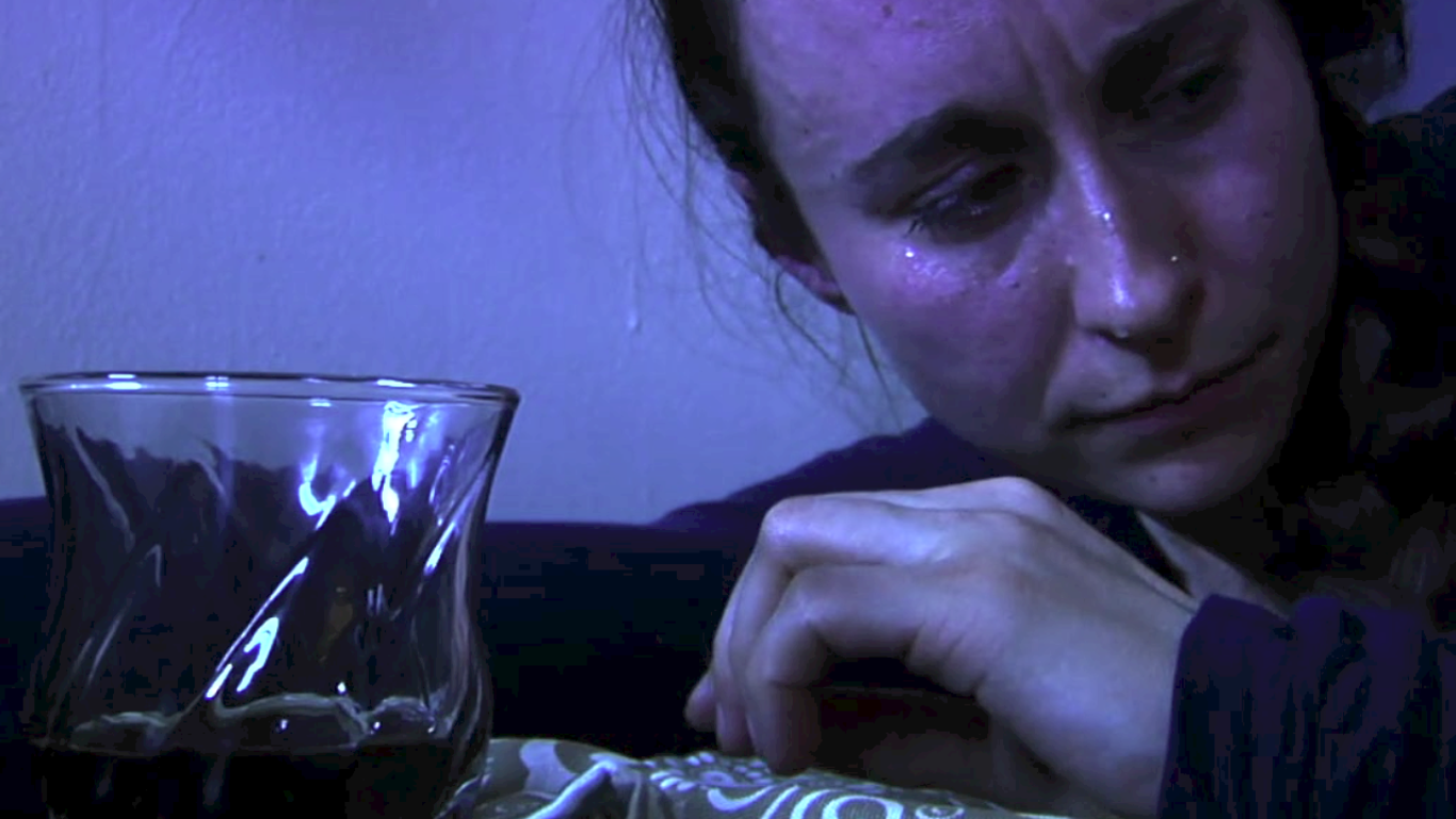 A woman sitting at a table with a glass of dark liquid, appearing sad or emotional.