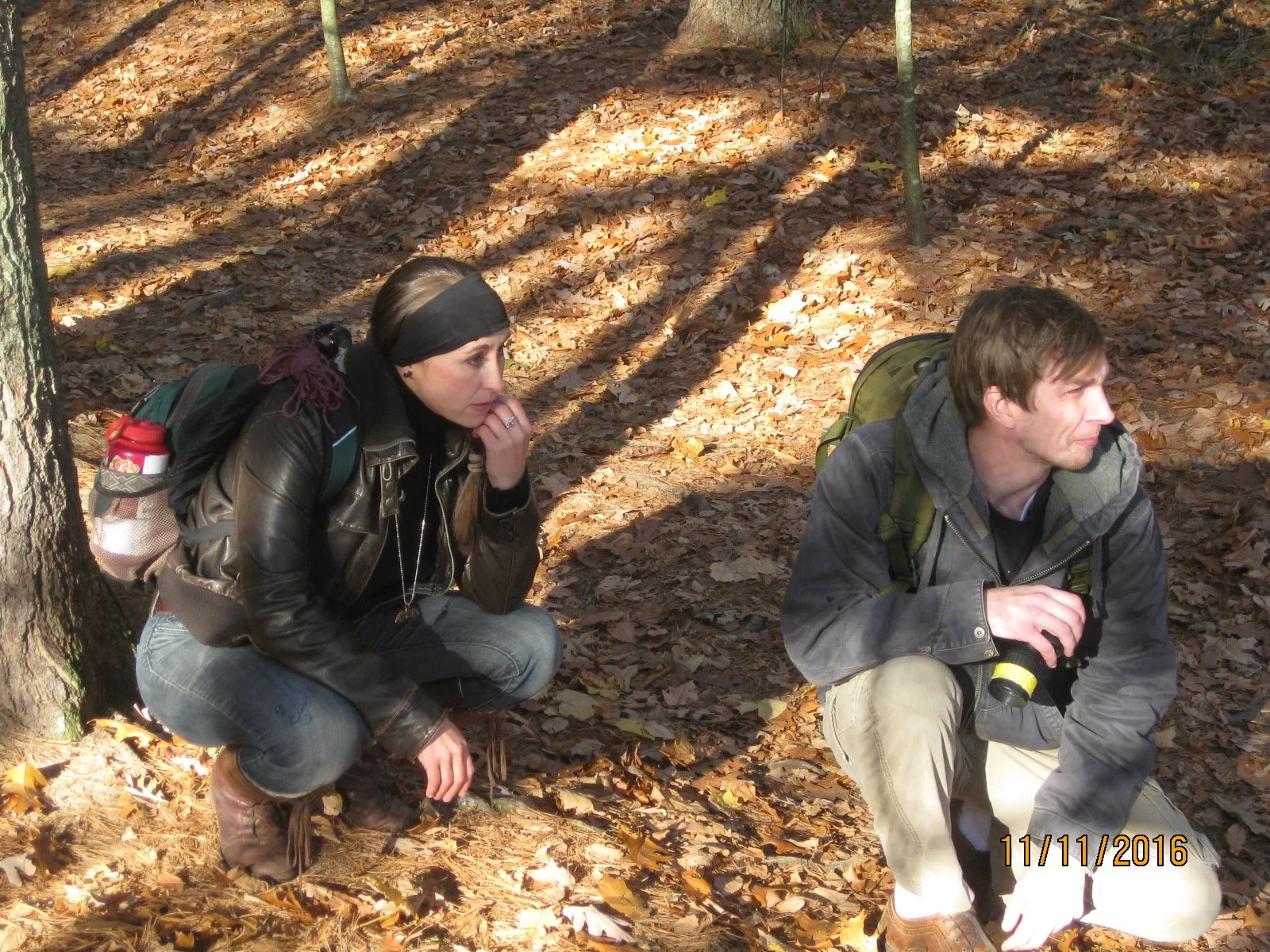 A woman and a man are crouching in a forest covered with fallen leaves. The woman is wearing a black leather jacket, a black headband, and has a backpack with a water bottle. Both are deep in thought, with the woman resting her chin on her hand and t
