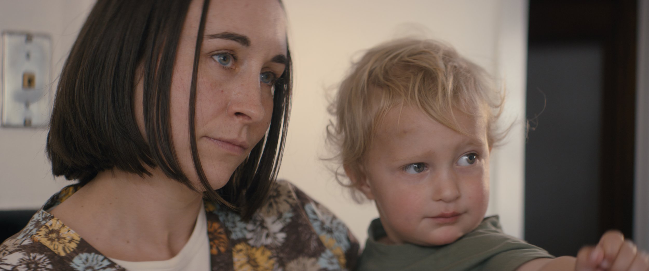 A woman with short dark hair and a young boy with curly blonde hair sitting together, both looking at something off-camera.