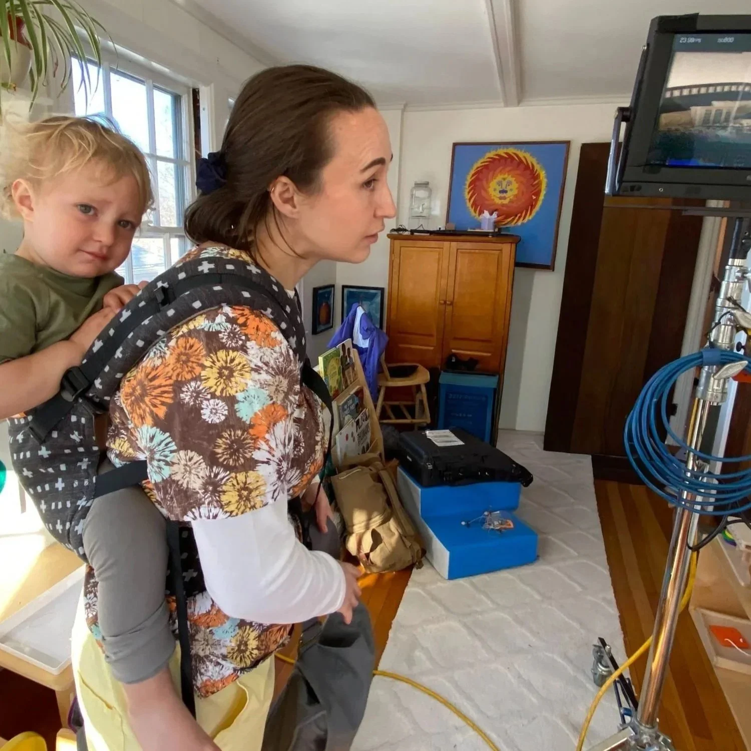 A woman carrying a young child on her back, both watching a monitor in a room with colorful art on the walls, and equipment including cables and boxes around them.