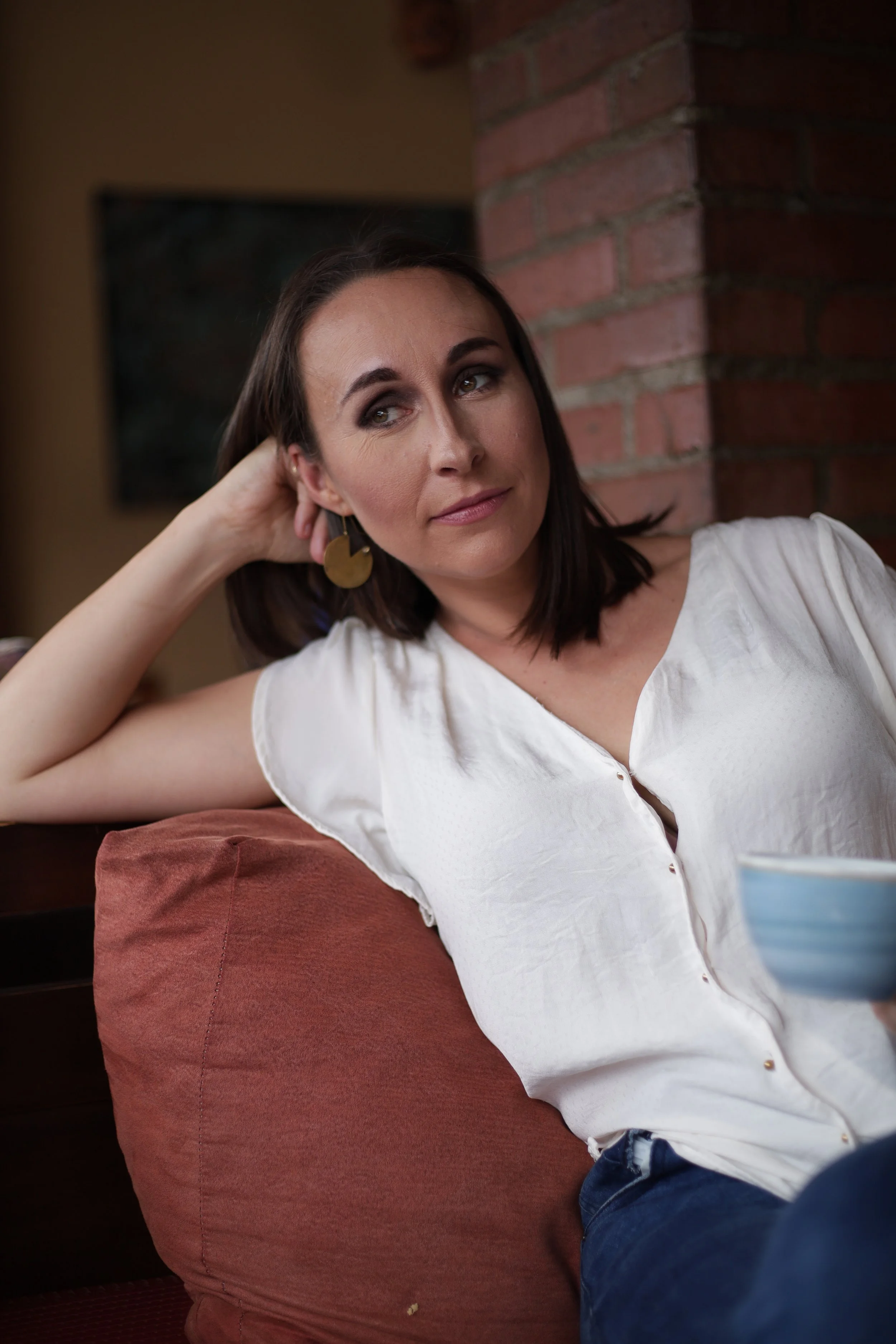 A woman with dark hair and gold earrings relaxing on a red cushion, leaning against a brick wall, wearing a white shirt and dark jeans, with a blue and white striped bowl in front of her.