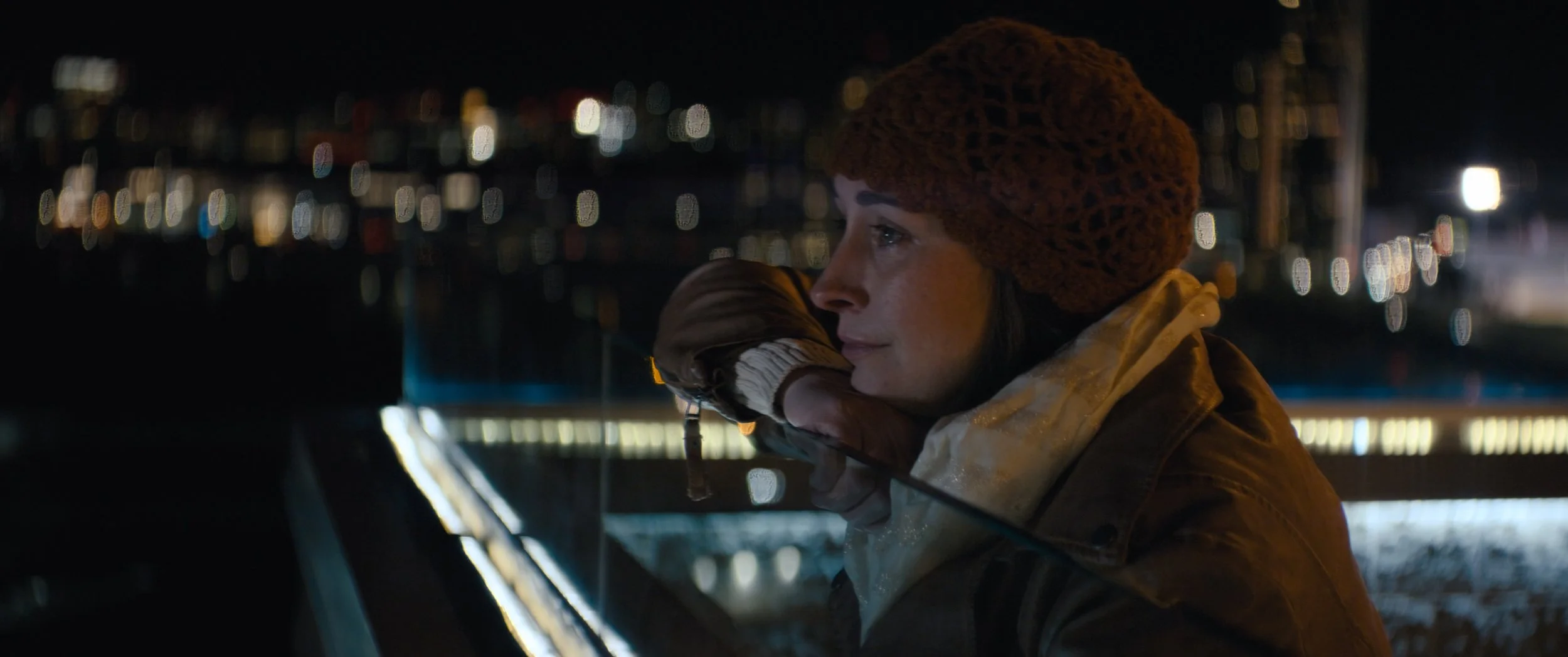A woman with short brown hair wearing a knitted brown hat and a brown jacket, leaning on a railing and gazing into the night sky with city lights blurred in the background.