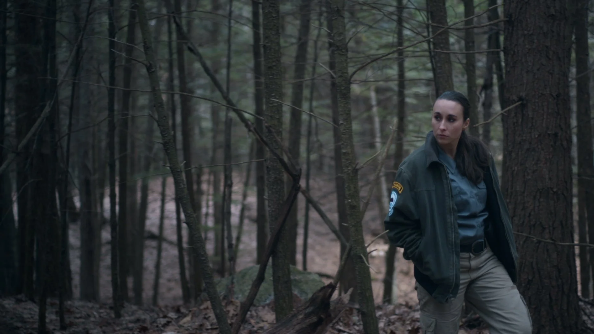 A woman in a sheriff uniform stands in a dense forest with tall trees and thin branches, looking to the side with a serious expression.