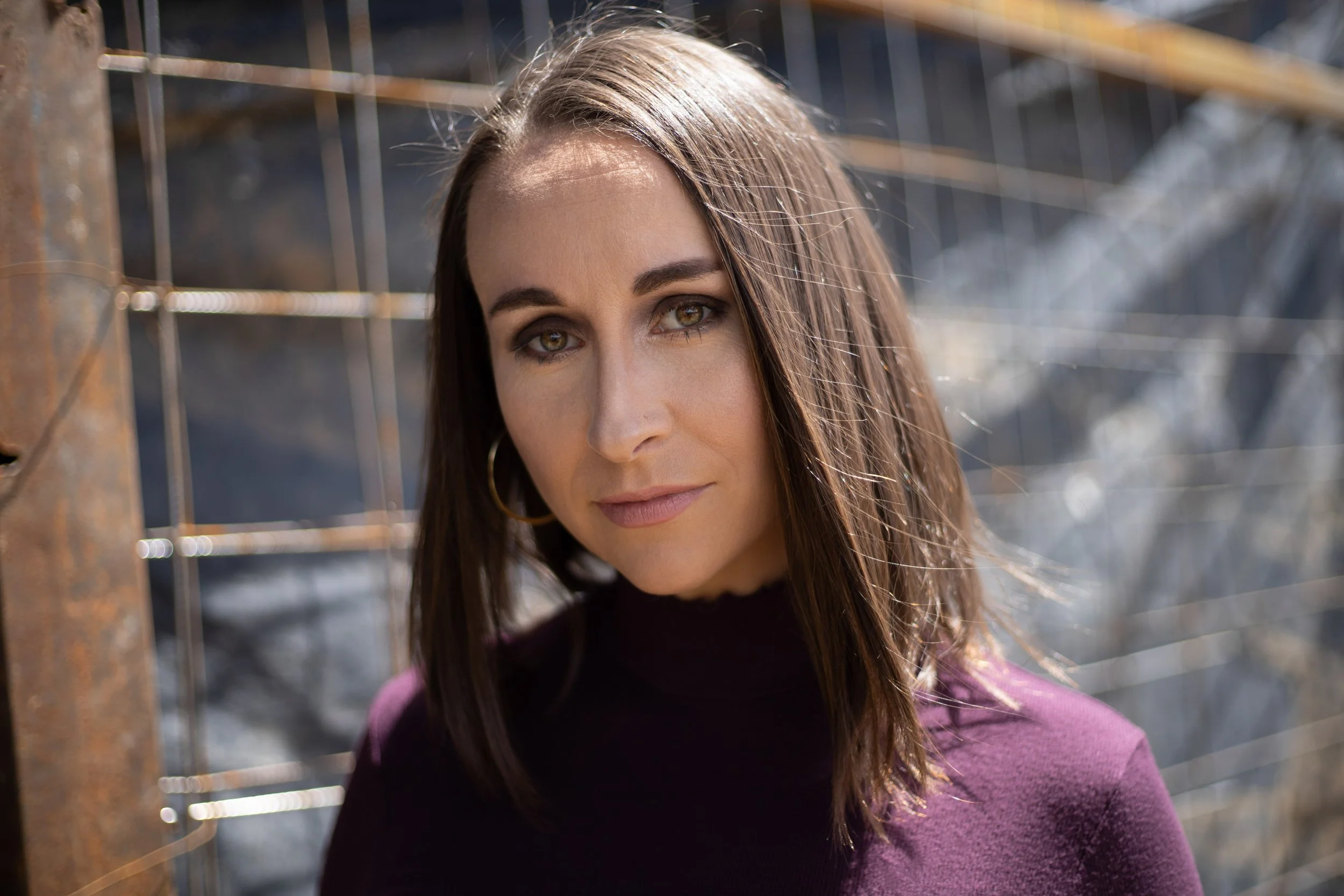 Close-up of a woman with brown hair and hazel eyes standing in front of a metal fence, wearing a purple sweater.