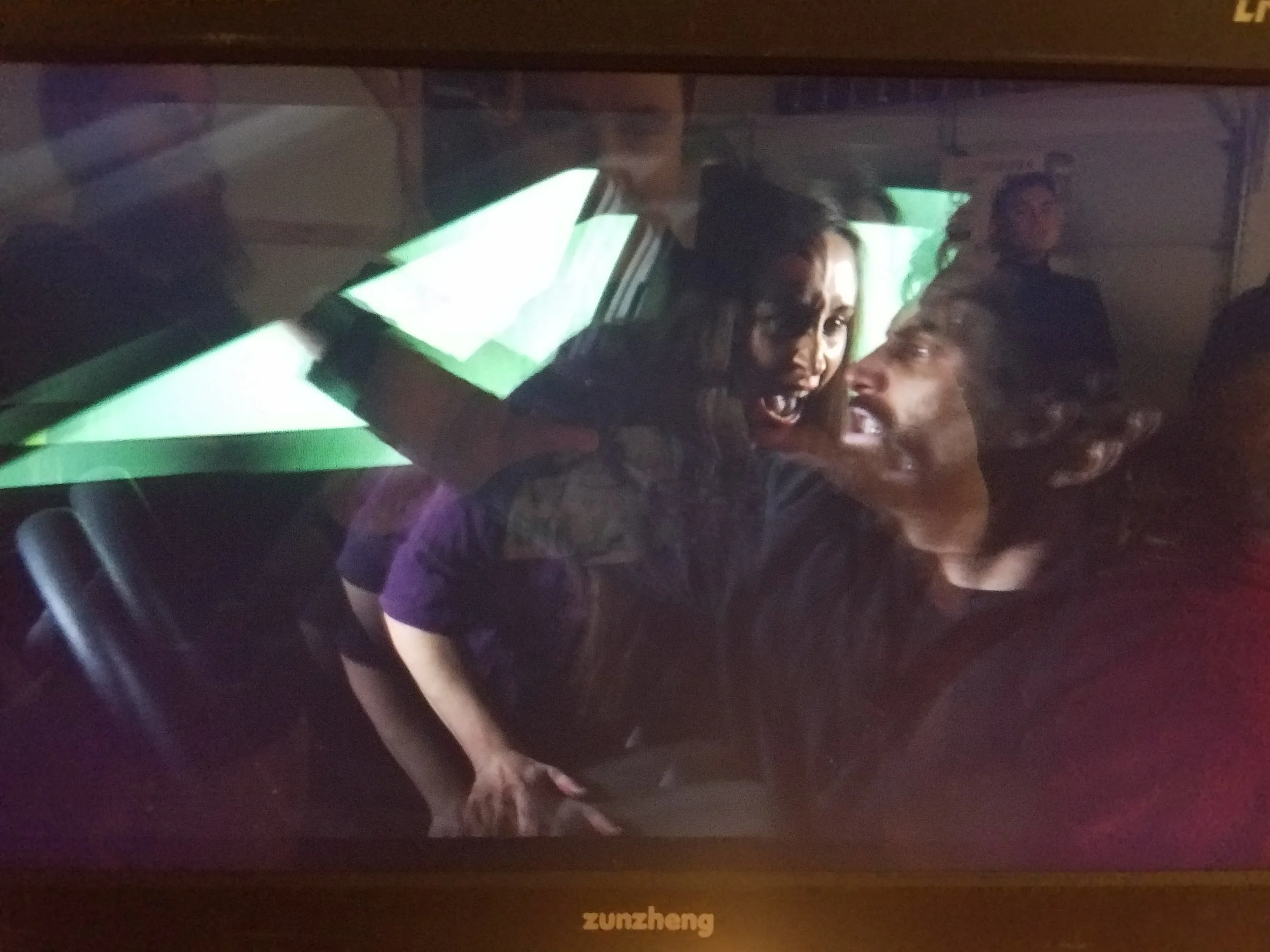 Two women taking a selfie in front of a computer screen displaying a green background, with other people sitting and working around them.