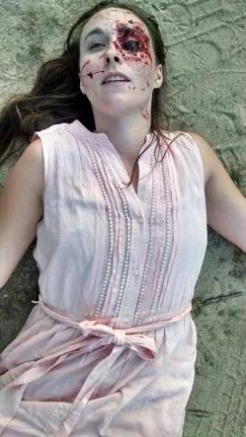 A girl lying on the ground with a serious injury or wound on her face, wearing a light pink dress, with long brown hair, on a sandy surface.