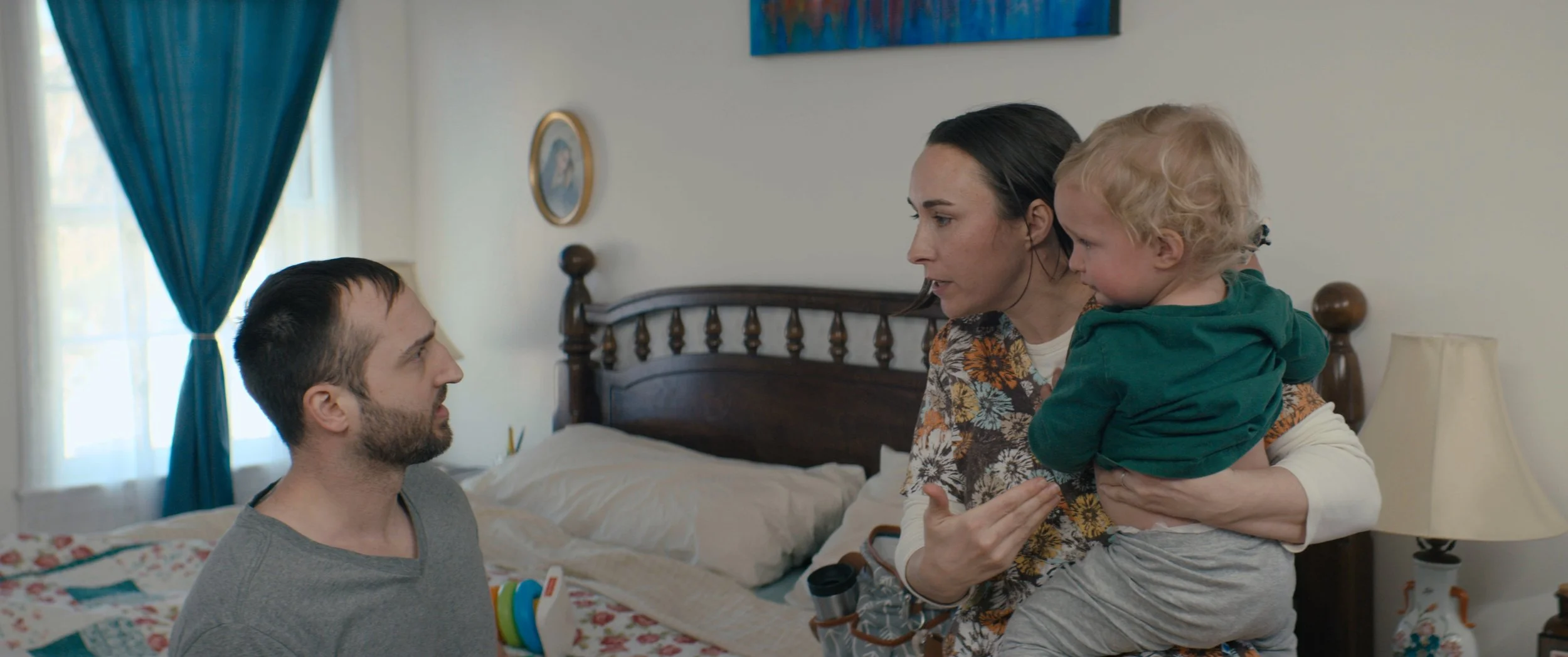 A woman holding a young child and talking to a man sitting on a bed in a bedroom.