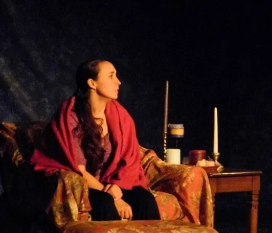 A woman with long dark hair, wearing a red shawl, sitting on a decorated sofa in a dark room with candles and candelabra on a small table nearby.
