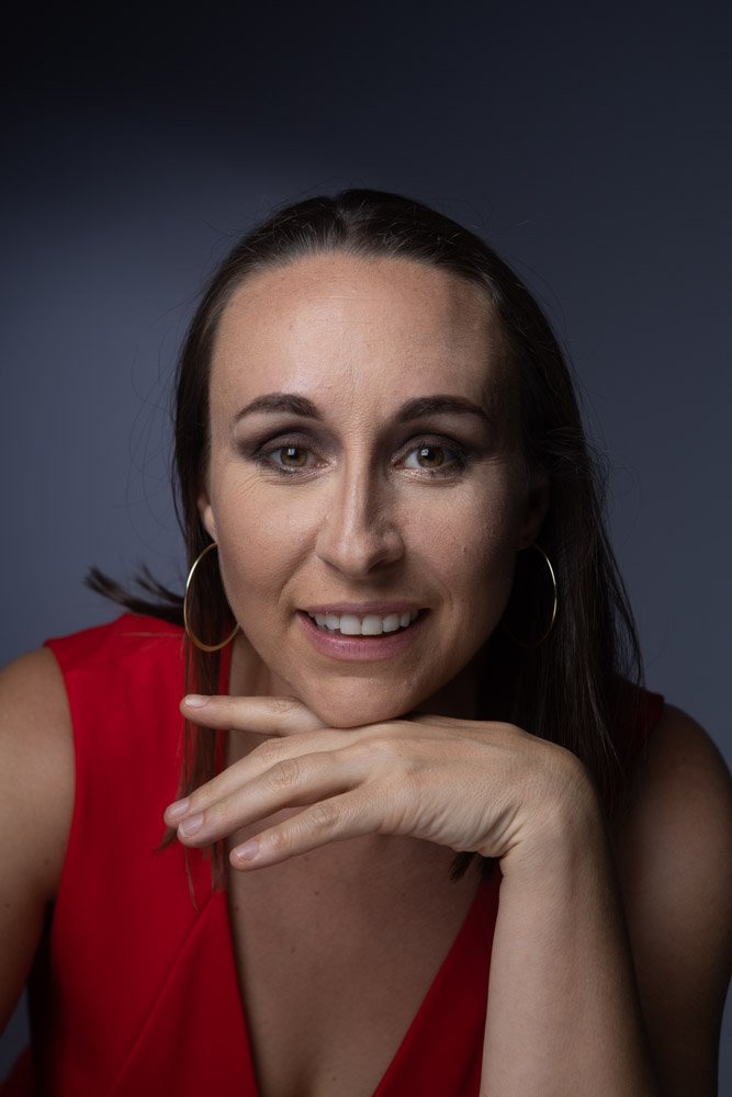 A woman with brown hair wearing a red sleeveless top and large hoop earrings, resting her chin on her hand, smiling at the camera with a dark background.