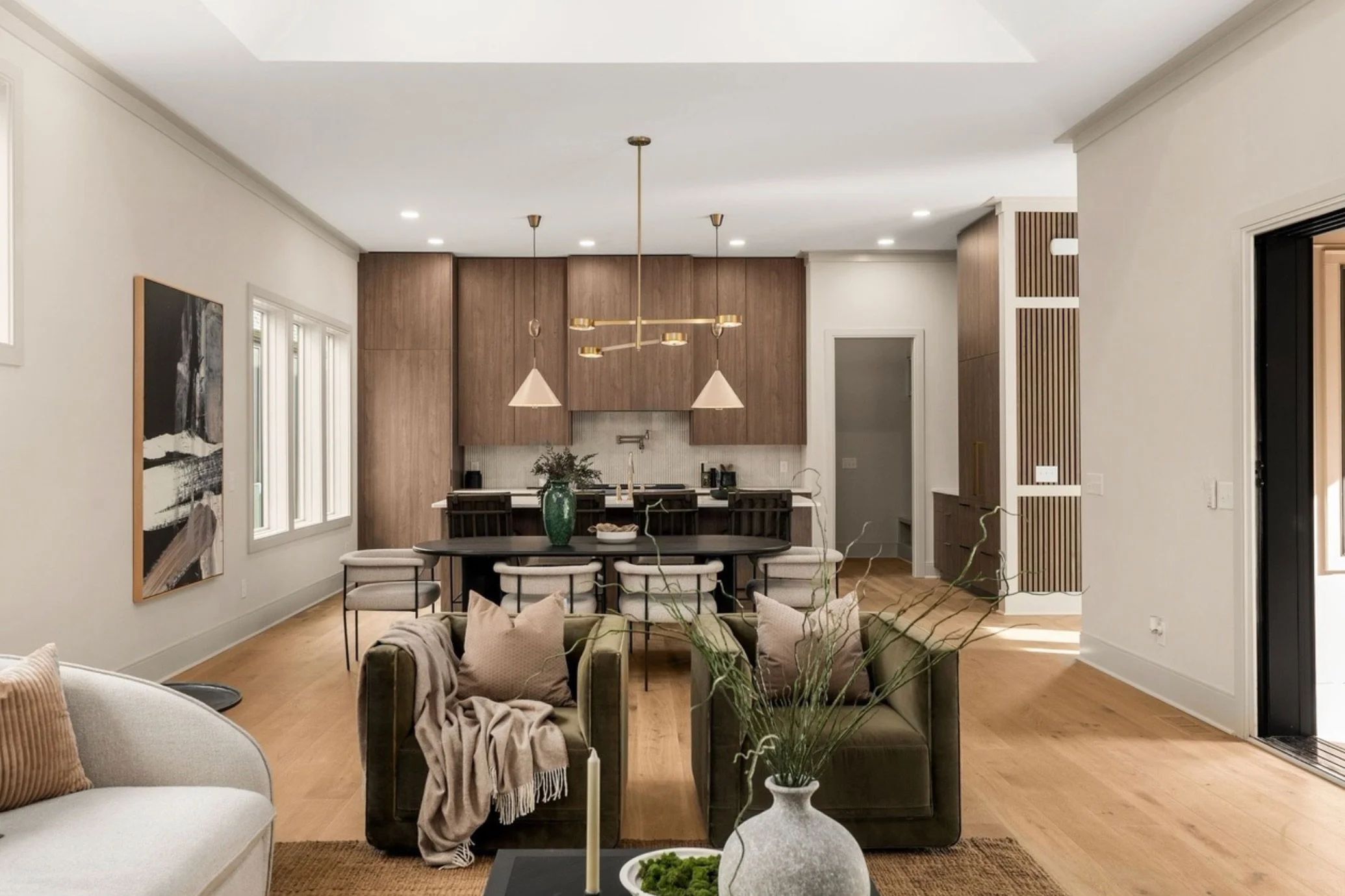 Modern living room with beige sofas, a black dining table surrounded by beige chairs, a large painting on the wall, wooden flooring, potted plants, and a kitchen area with wooden cabinets and white walls.