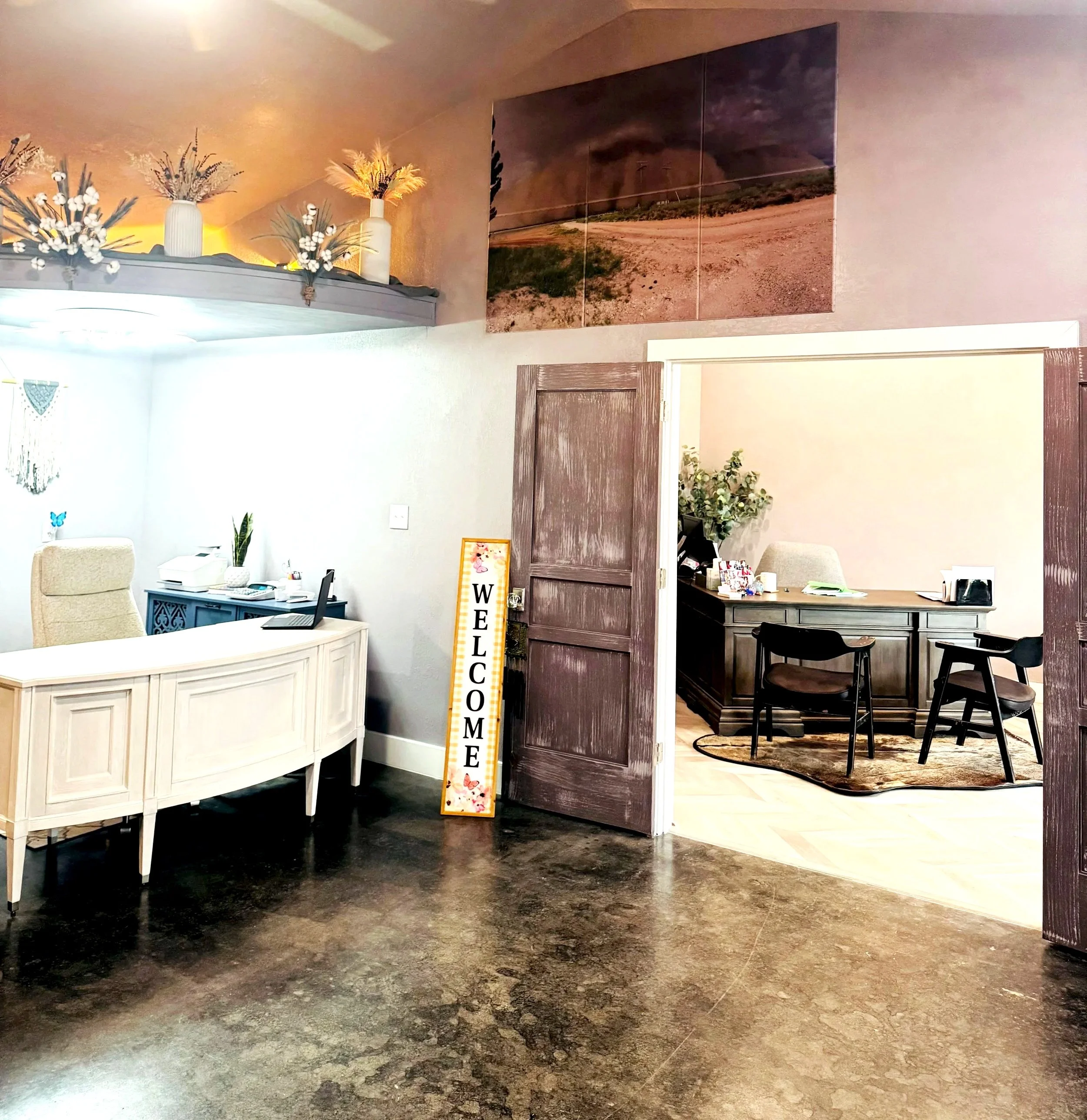 Office space with a reception desk, decorative vases with artificial flowers on a high shelf, a framed picture, and a sign saying 'Welcome'. There is a wooden door, and part of a meeting area with chairs and a rug is visible.