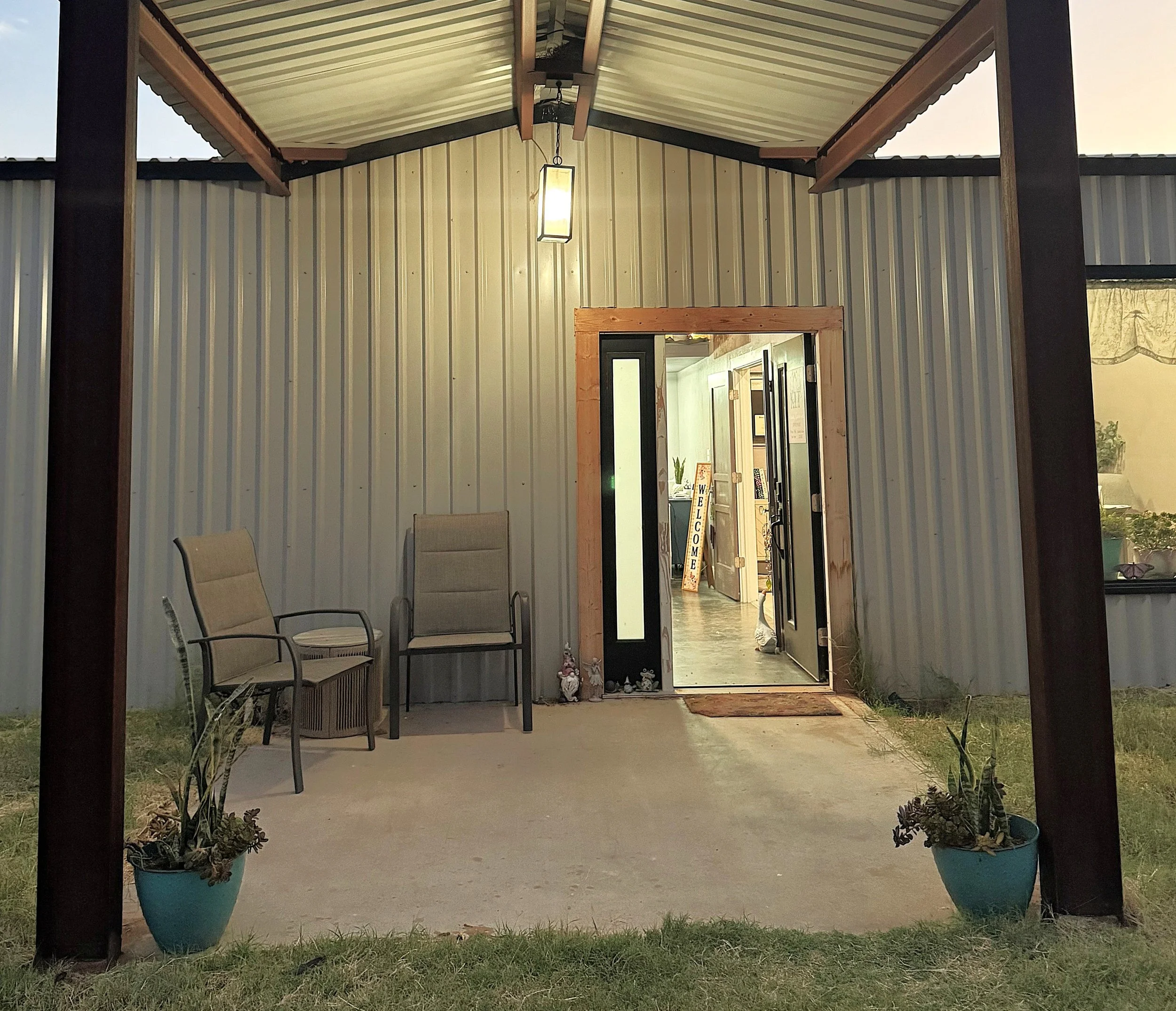 The image shows a covered porch area with two chairs and potted plants at the front. The porch has a metal and wood structure, with a hanging light fixture above the entrance door. Inside, there is a sign that says 'WELCOME' standing next to the doorway. The view looks into a well-lit room with some furniture and decorations visible.