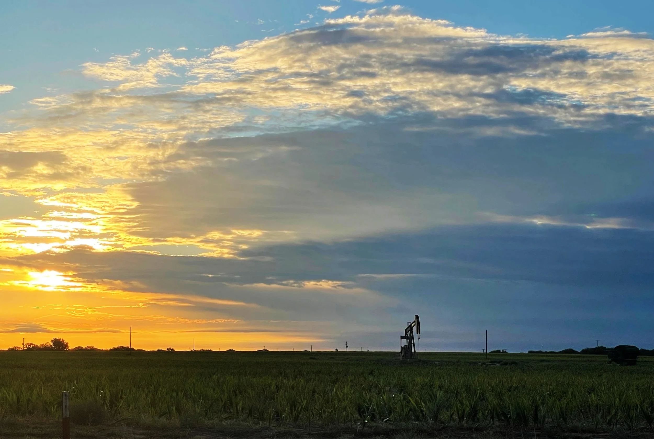 A sunrise or sunset over a flat rural landscape with green fields, a few scattered trees, some electrical towers, and a pumpjack in the distance, with a partly cloudy sky.