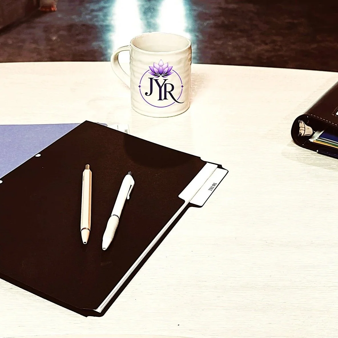 A white table with a black folder, two white pens, a white notebook, a brown wallet with several colored cards inside, and a mug with a purple lotus flower logo and the letters 'JYR' on it.