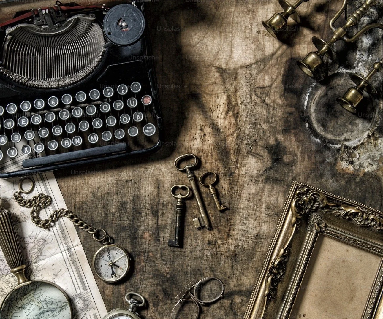 Vintage black typewriter, three old keys, the frame of an empty ornate picture frame, antique pocket watch, pair of glasses, brass keys, old map, and brass candle holders on a rustic wooden table.