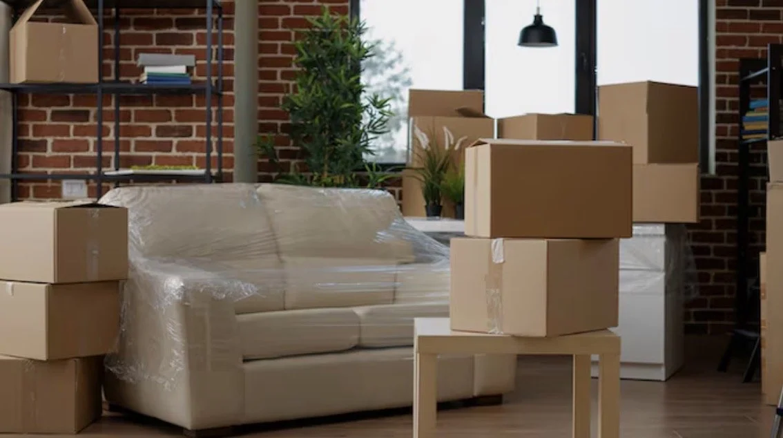 Living room with cardboard boxes, some on a small table, a sofa covered in plastic, brick wall, bookshelf, and large window with plants.