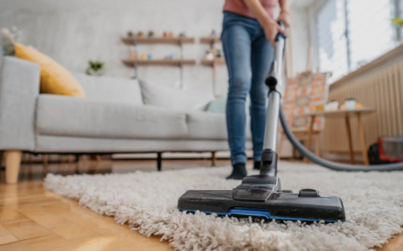 Person vacuuming a beige carpet in a living room with a gray sofa, yellow pillow, and wooden shelves with decor in the background.