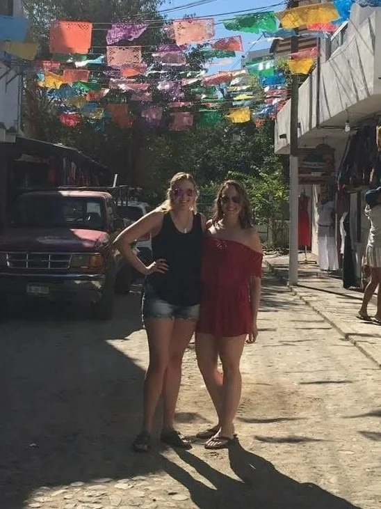 Two women standing close together on a sunlit street, under colorful paper decorations hanging above, with shops and a vehicle in the background.