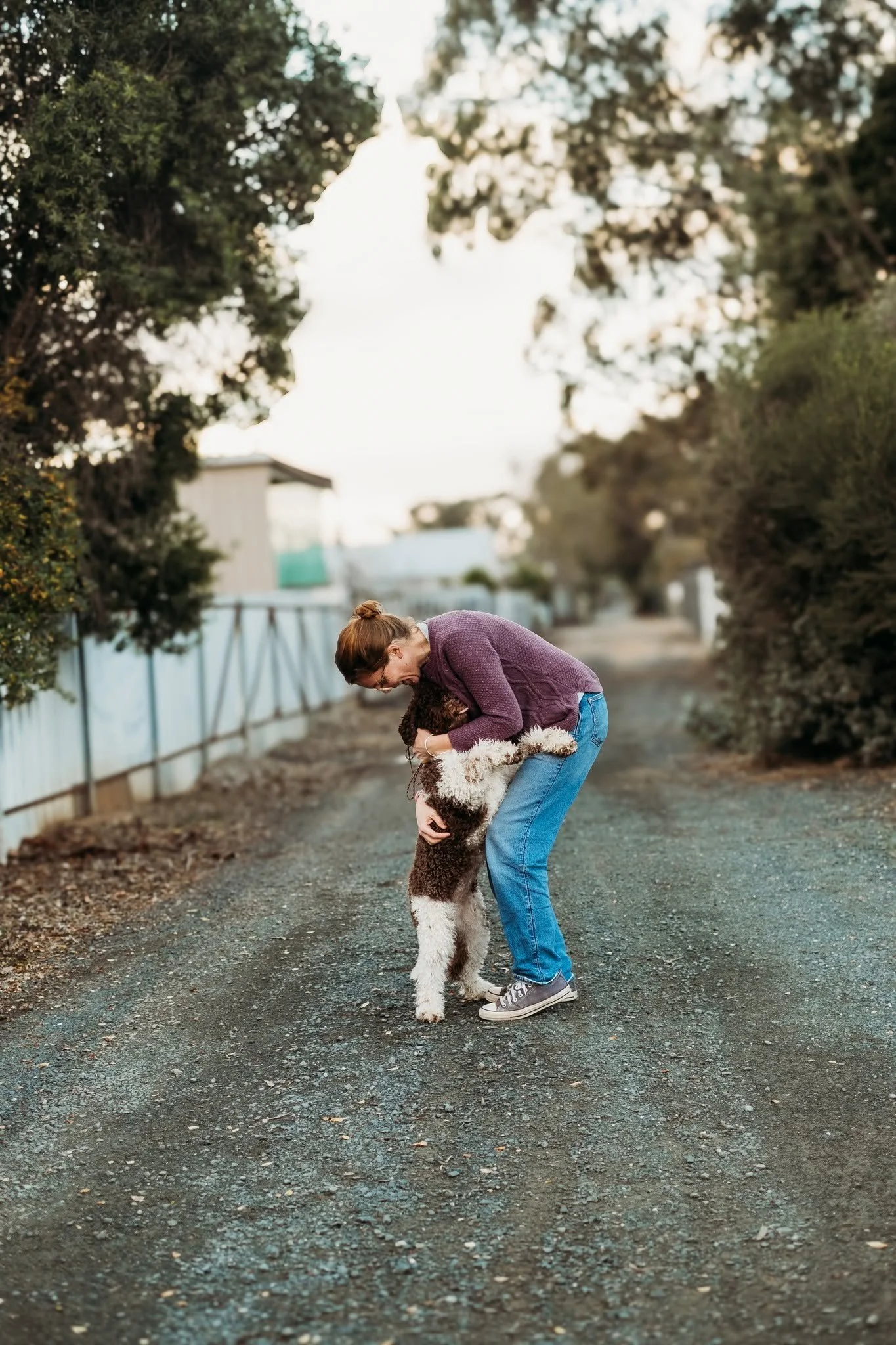A woman with glasses and a bun, wearing a purple sweater and jeans, bending down and hugging a large, fluffy dog on a gravel path in a suburban or rural outdoor setting during late afternoon or early evening.