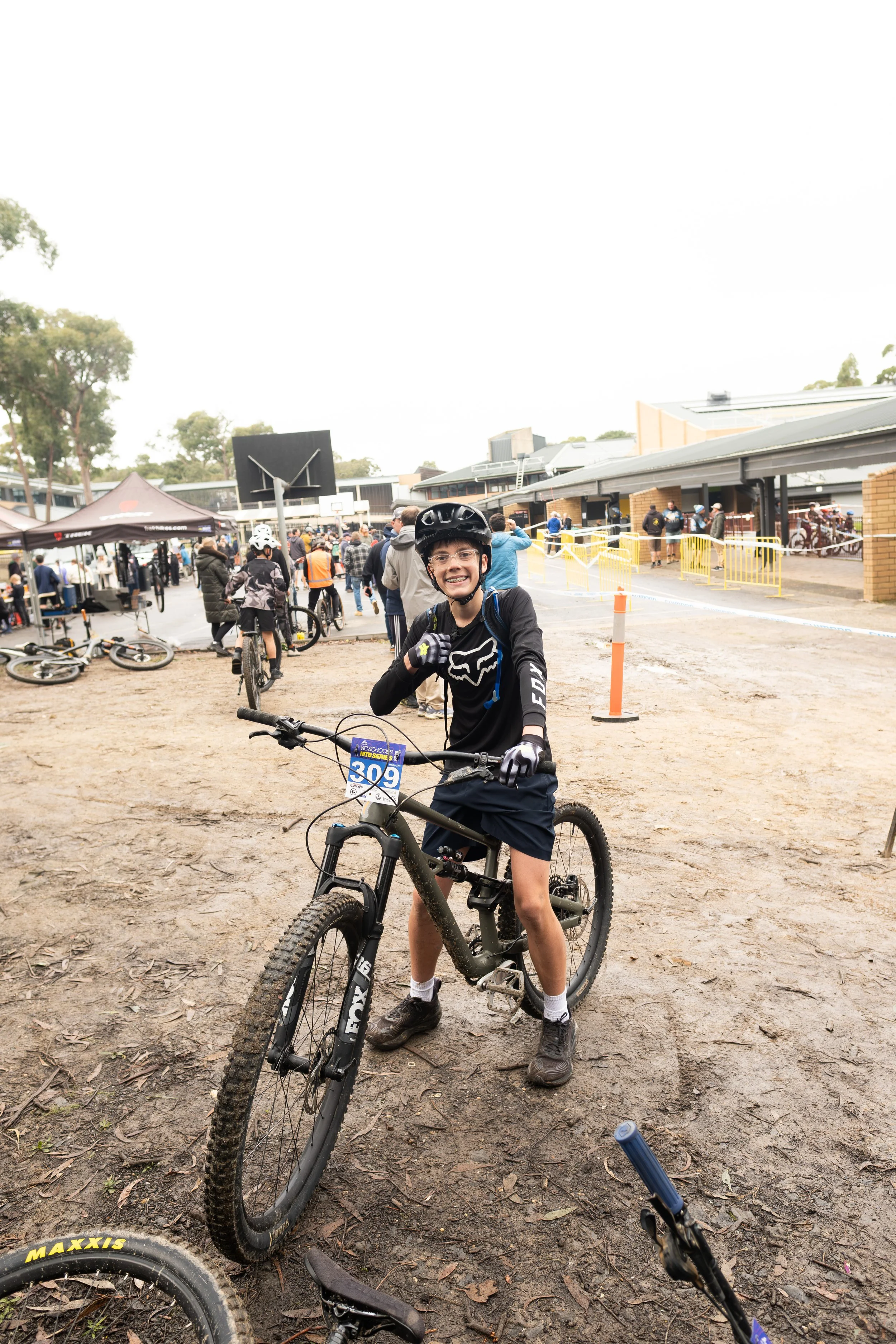 A young boy standing with a mountain bike at an outdoor event, smiling, wearing a black helmet, black long sleeve shirt, black shorts, and black shoes, in a crowded area with other cyclists, tents, and a school building in the background.