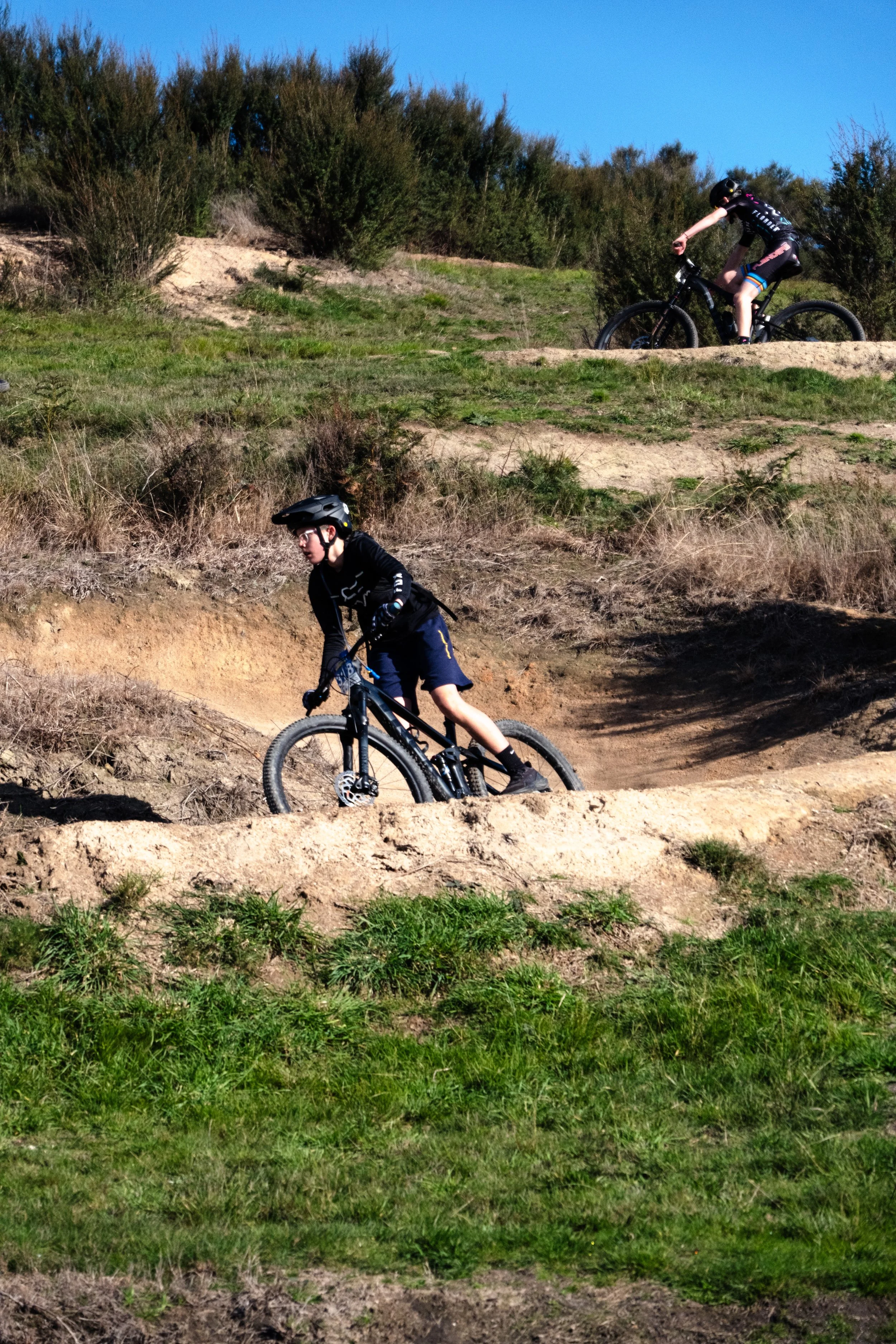 Two children riding mountain bikes on a dirt trail in a hilly, grassy area near bushes and trees with clear blue sky.