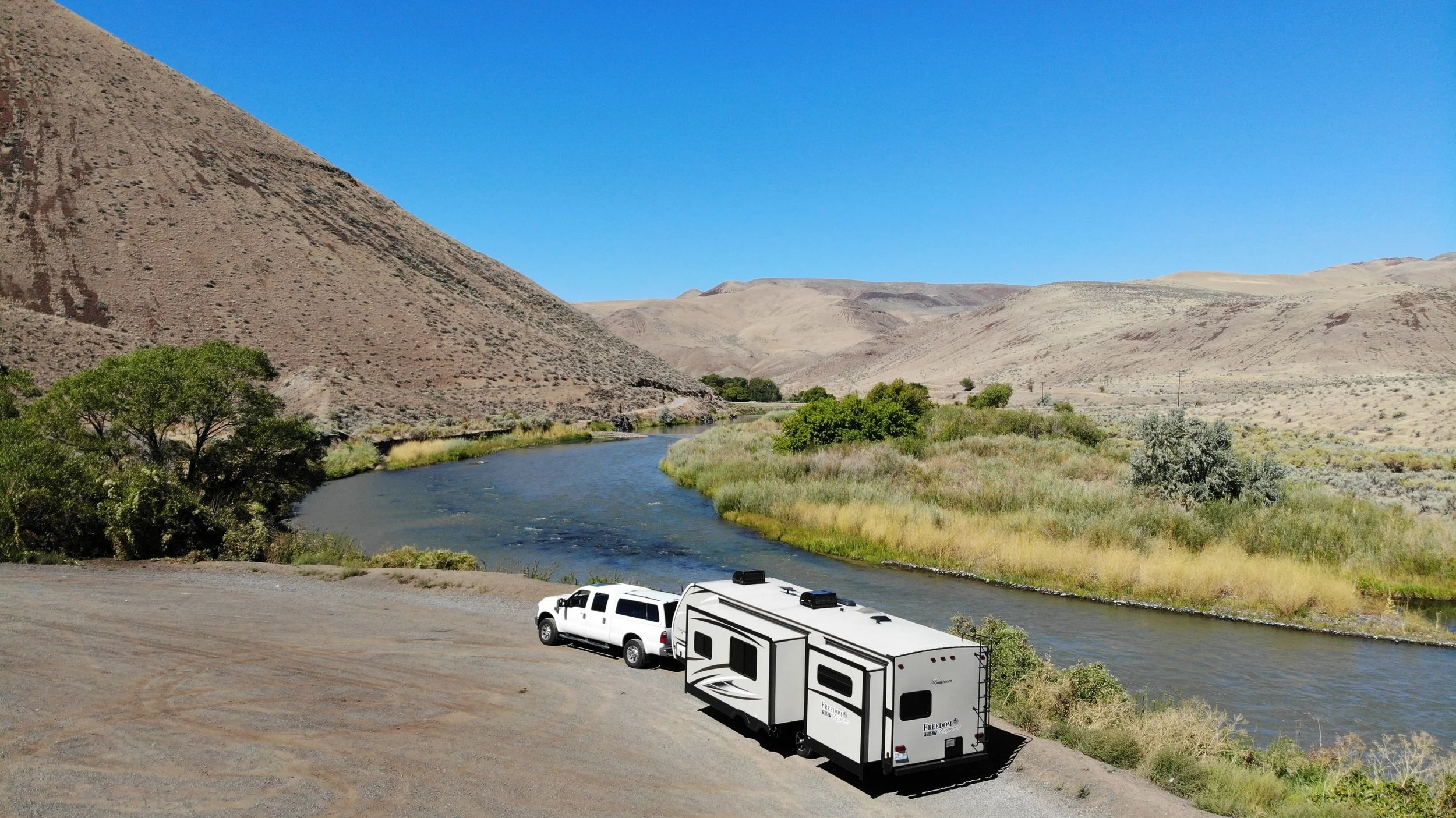 A truck and travel trailer parked beside a river in dry, grassy hills under a clear blue sky.
