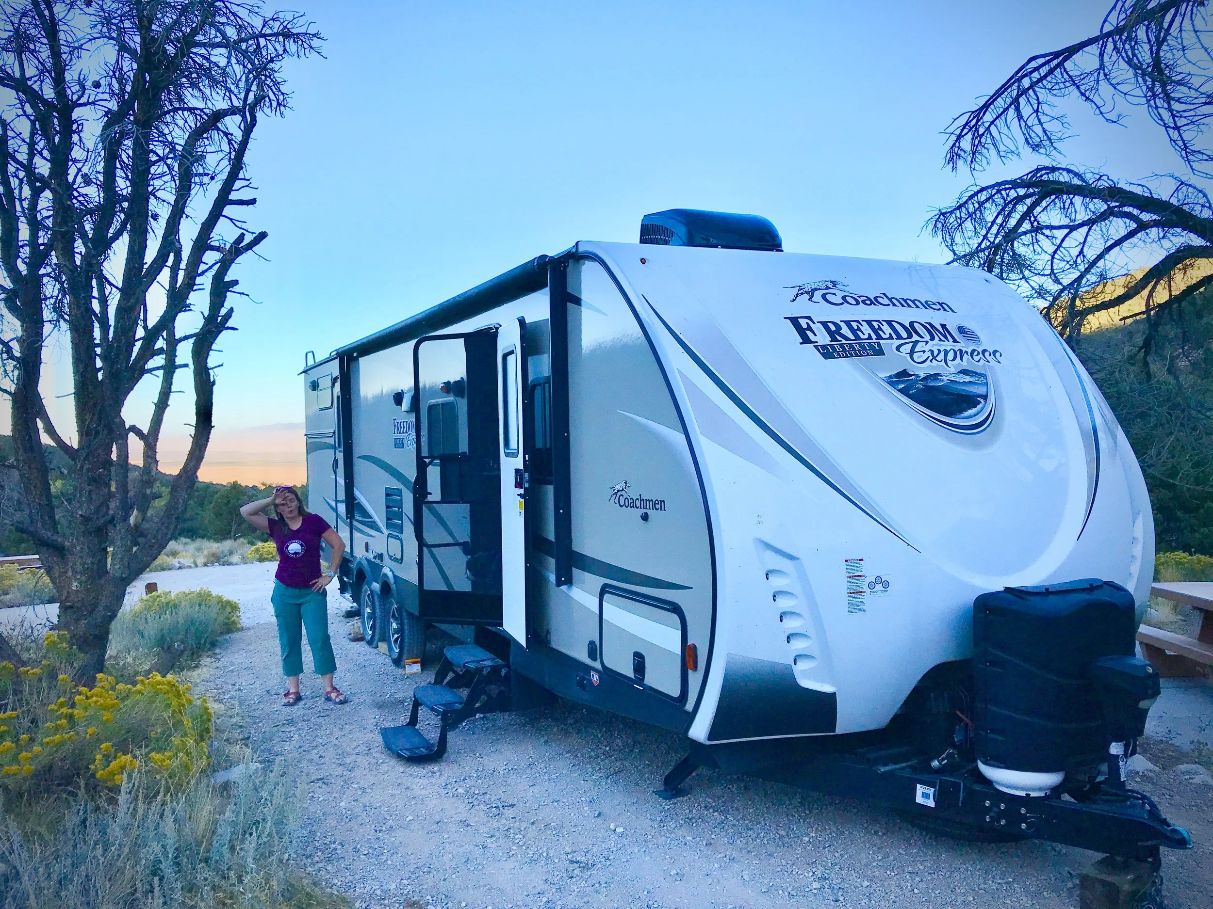 A woman stands beside a Coachmen Freedom Express travel trailer parked at a riverside campsite at dusk.