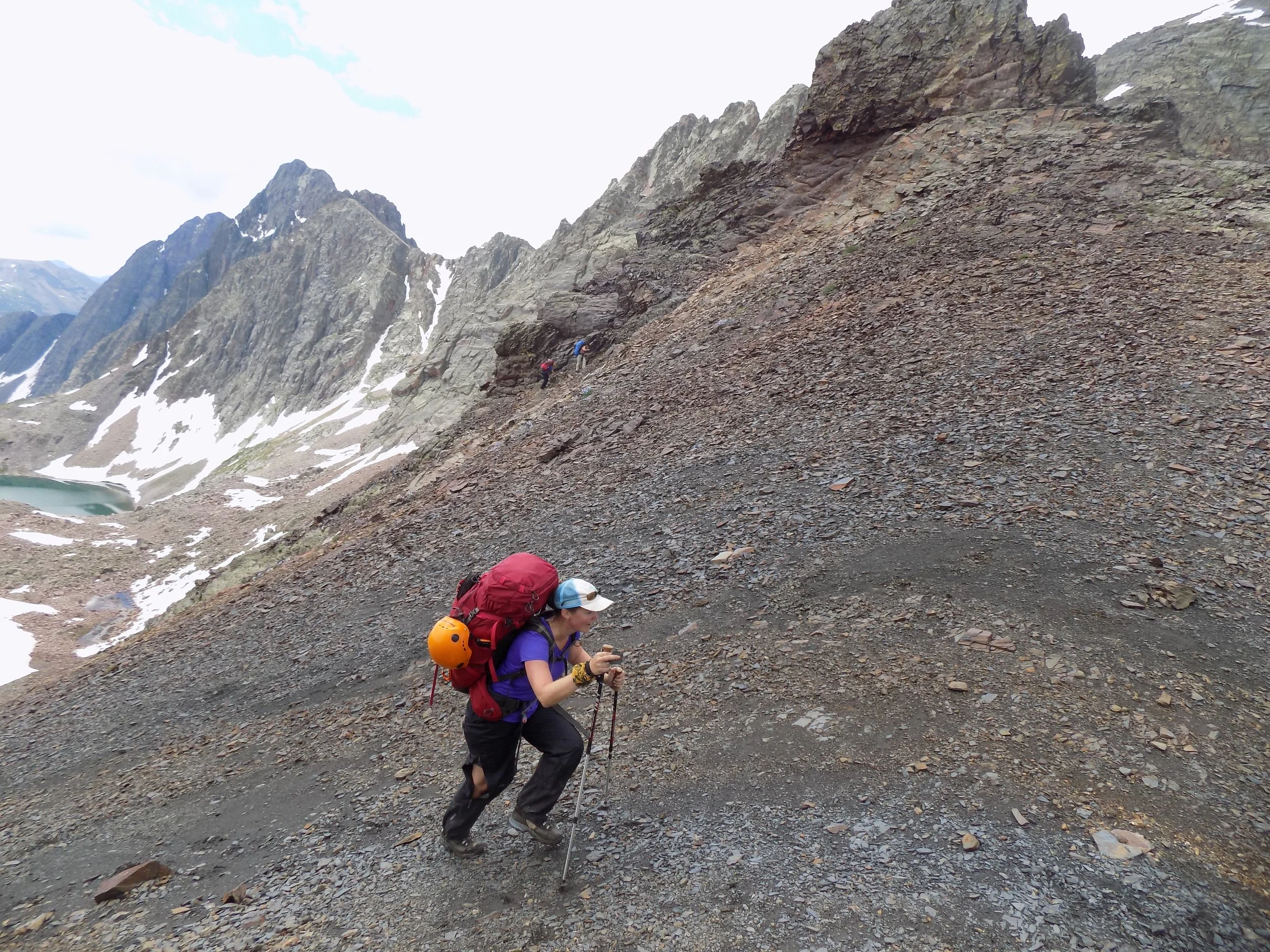 A hiker with an orange backpack climbing a rocky scree slope below patches of snow.