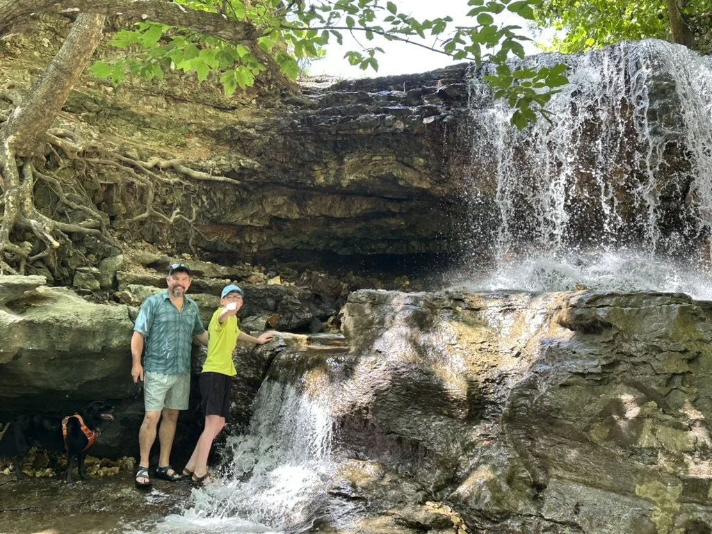 A man, boy, and their dog stand at the base of a small rock bluff with a waterfall cascading over.