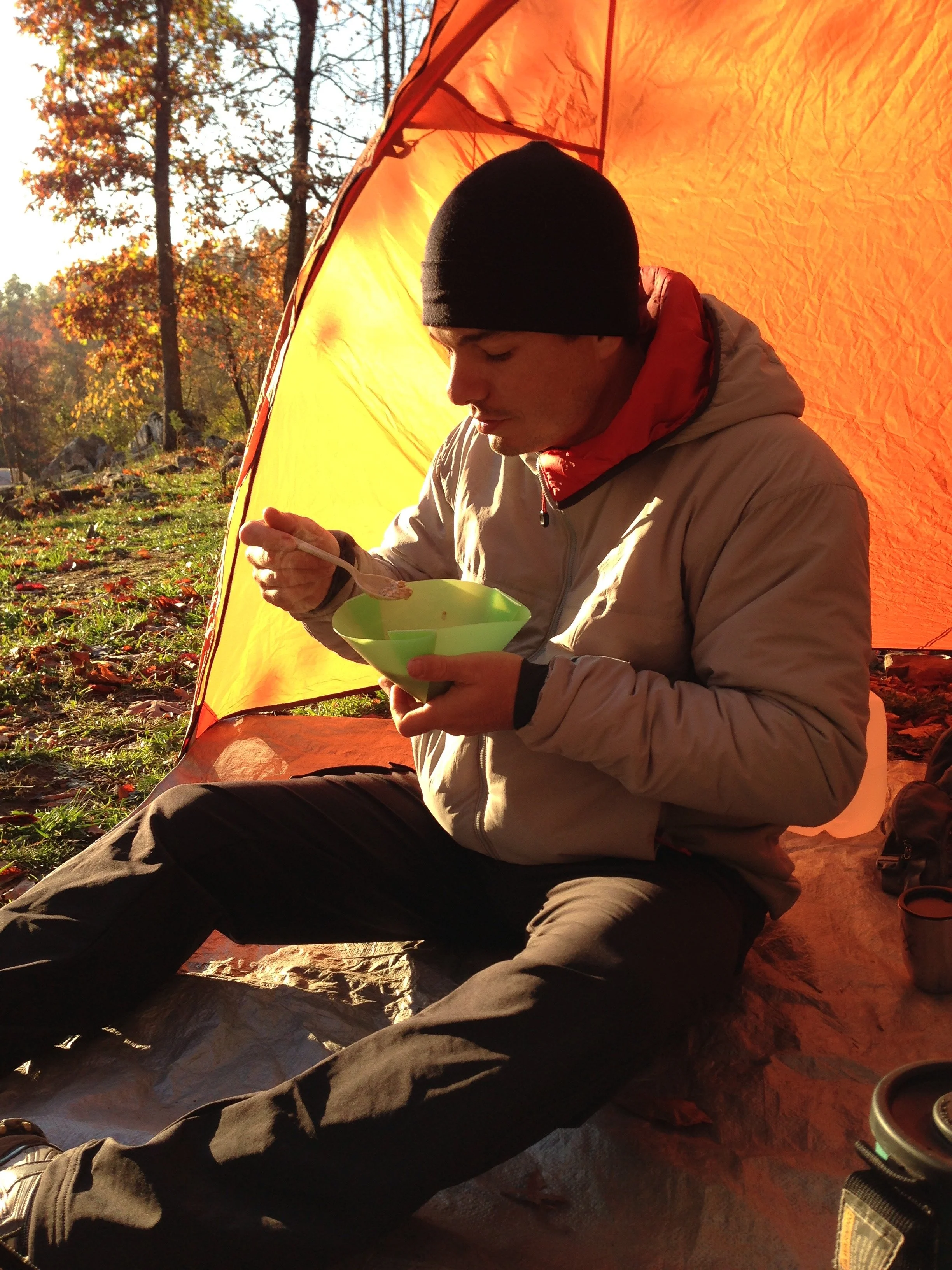 A person in a tan jacket sitting in the doorway of an orange tent eating from a green bowl, autumn trees behind.