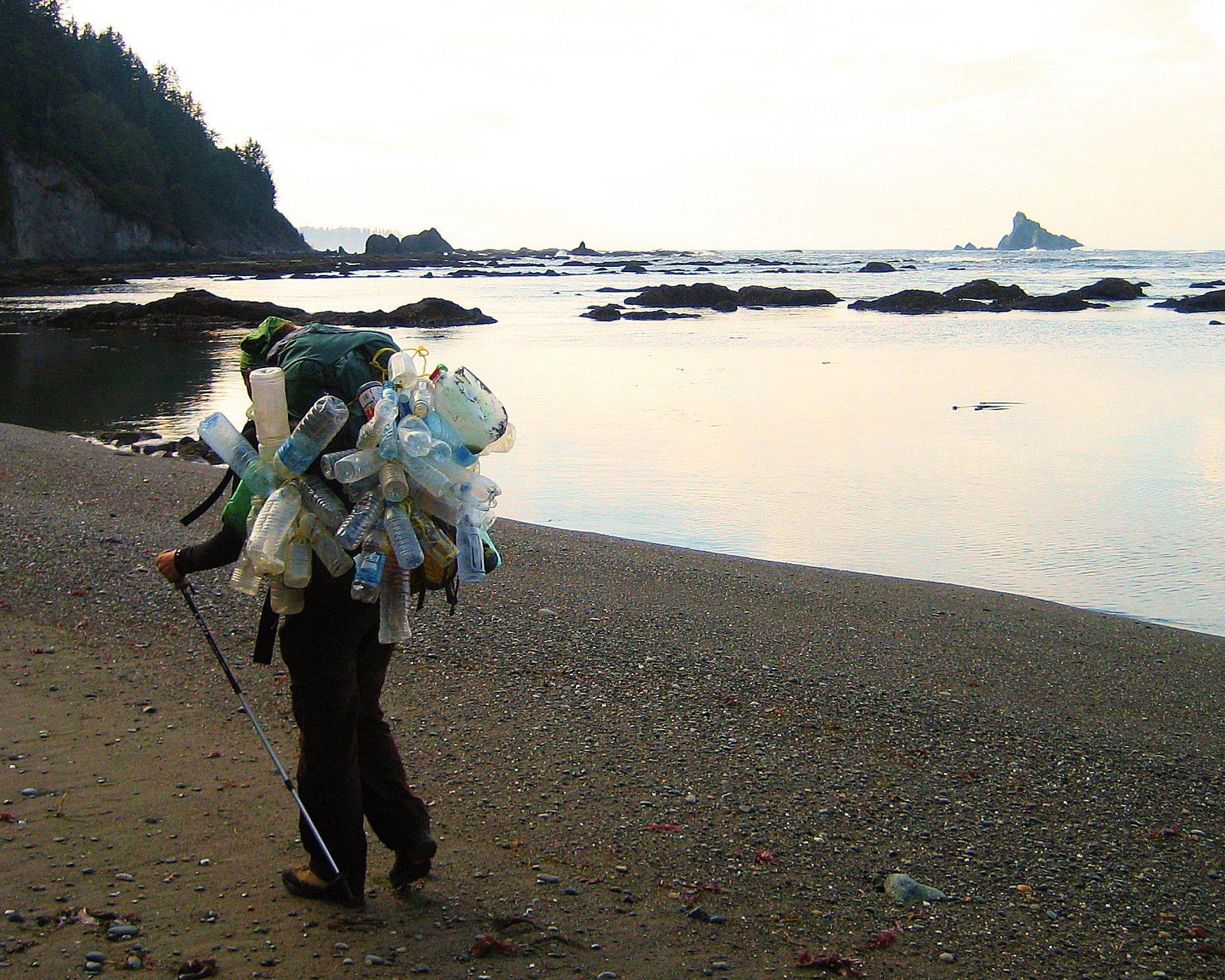 A backpack covered in dozens of collected plastic bottles resting on a rocky Pacific beach at golden hour, sea stacks and forested cliffs beyond.