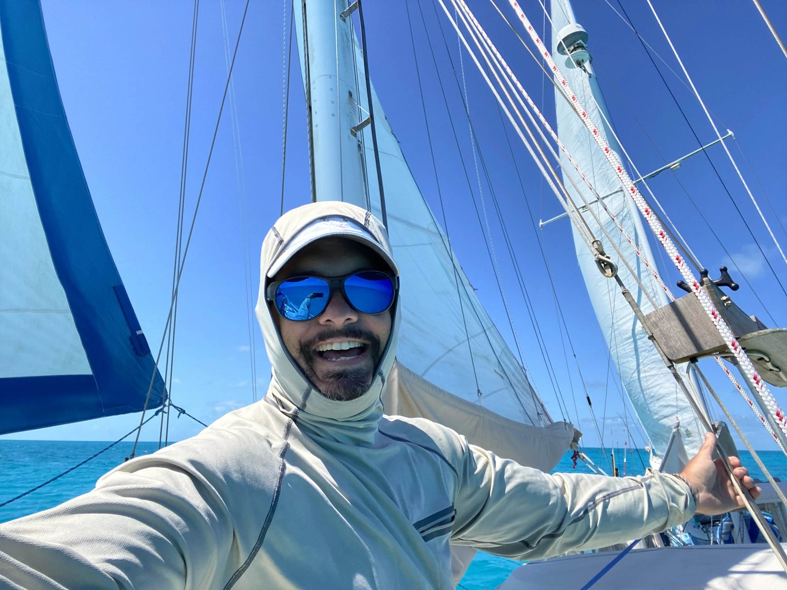 A smiling sailor in a sun hoodie and sunglasses at the helm beneath full sails.