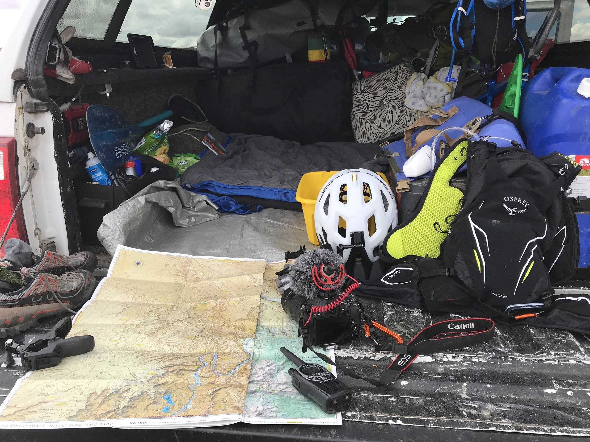 Outdoor gear spread out in the back of a vehicle: a bike helmet, backpacks, shoes, and a folded paper map.